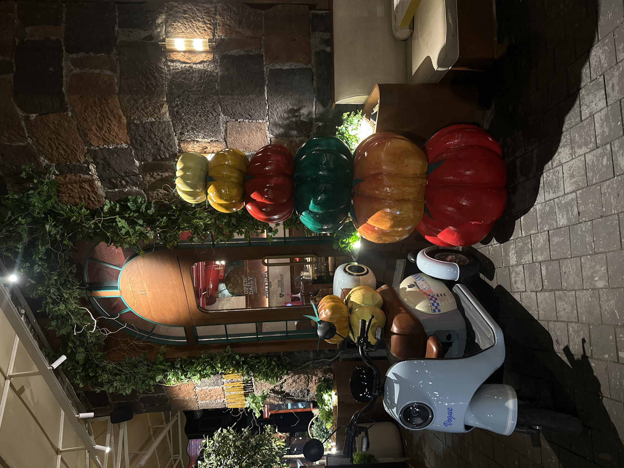 The image is a nighttime shot of the interior of a restaurant, or a decorative setup outside one, located in Yerevan, Armenia.
A cobblestone or brick wall with artificial plants adorns the top. In front of this are a series of colorfully painted pumpkin-shaped decorations: green, yellow, red, and orange.
Below this decorative layer is a restaurant window with a display of pasta behind it, indicating Italian cuisine. Above, foliage and a decorative arch add to the ambiance.
A light blue motor scooter bearing the logo "Vogue" is parked in the foreground and faces the entrance, partly obscuring the view of the restaurant's interior. The scooter is flanked by colorful gourds and pumpkins, including some with added features like a bee attached to them, contributing to a decorative, autumn-themed display.