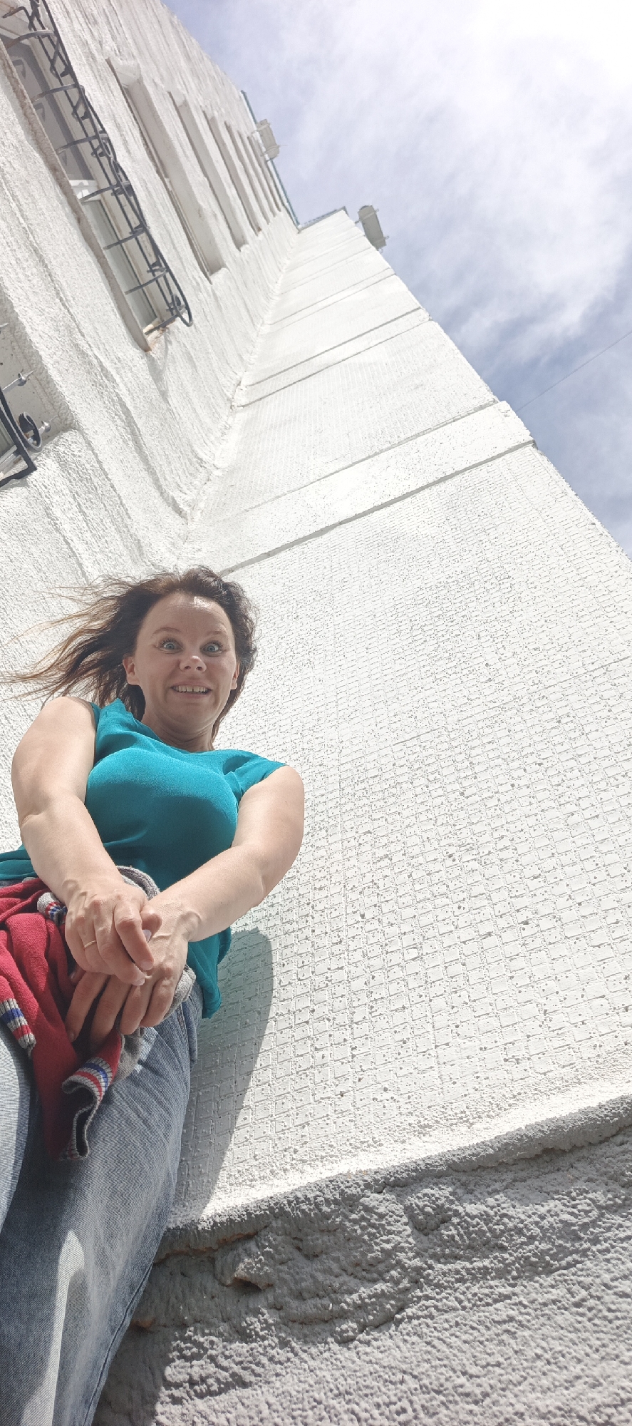 A low-angle shot captures a woman standing against a tall, white, textured building. She is looking upwards at the camera with a surprised and playful expression, her eyes wide and mouth open in a smile. Her brown hair is windswept, suggesting it is a breezy day. She is wearing a teal-colored sleeveless top and blue jeans. A red garment with striped edges is draped over her arms. The building behind her is a stark white, with a series of windows that have dark metal bars over them. The building's texture is rough and uneven, and a grey, rough, textured ledge runs along the bottom of the frame. The sky above is a pale blue with scattered white clouds, indicating daylight. The overall impression is one of a candid, spontaneous moment captured outdoors in Chaikovsky, Russia.