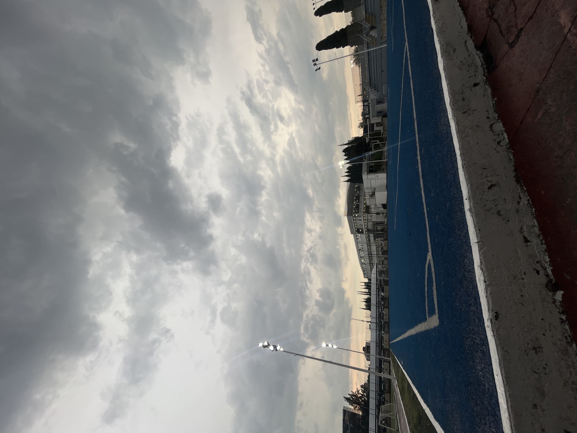 The image depicts an outdoor athletic track under a cloudy sky. The track is a combination of blue and red surfaces, with white lines marking the lanes and boundaries. Stadium lights are visible, and a building is in the background. The location is Unknown, Unknown.