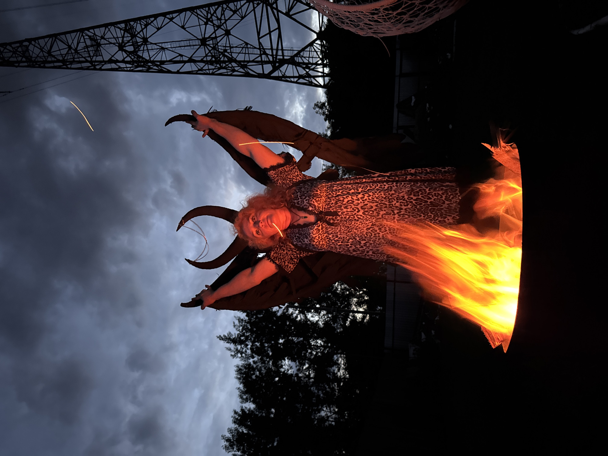 A person wearing a leopard-print dress and large, dark wings stands with their arms raised in front of a fire pit. Two large, curved horns are attached to their head. The person is holding a glowing stick or cigarette in their mouth. The scene takes place outdoors at dusk, with a cloudy sky and a large industrial tower in the background. Trees and a fence are visible behind the person. The overall atmosphere is dramatic and somewhat menacing. The location is the city of Chaikovsky, Russia.