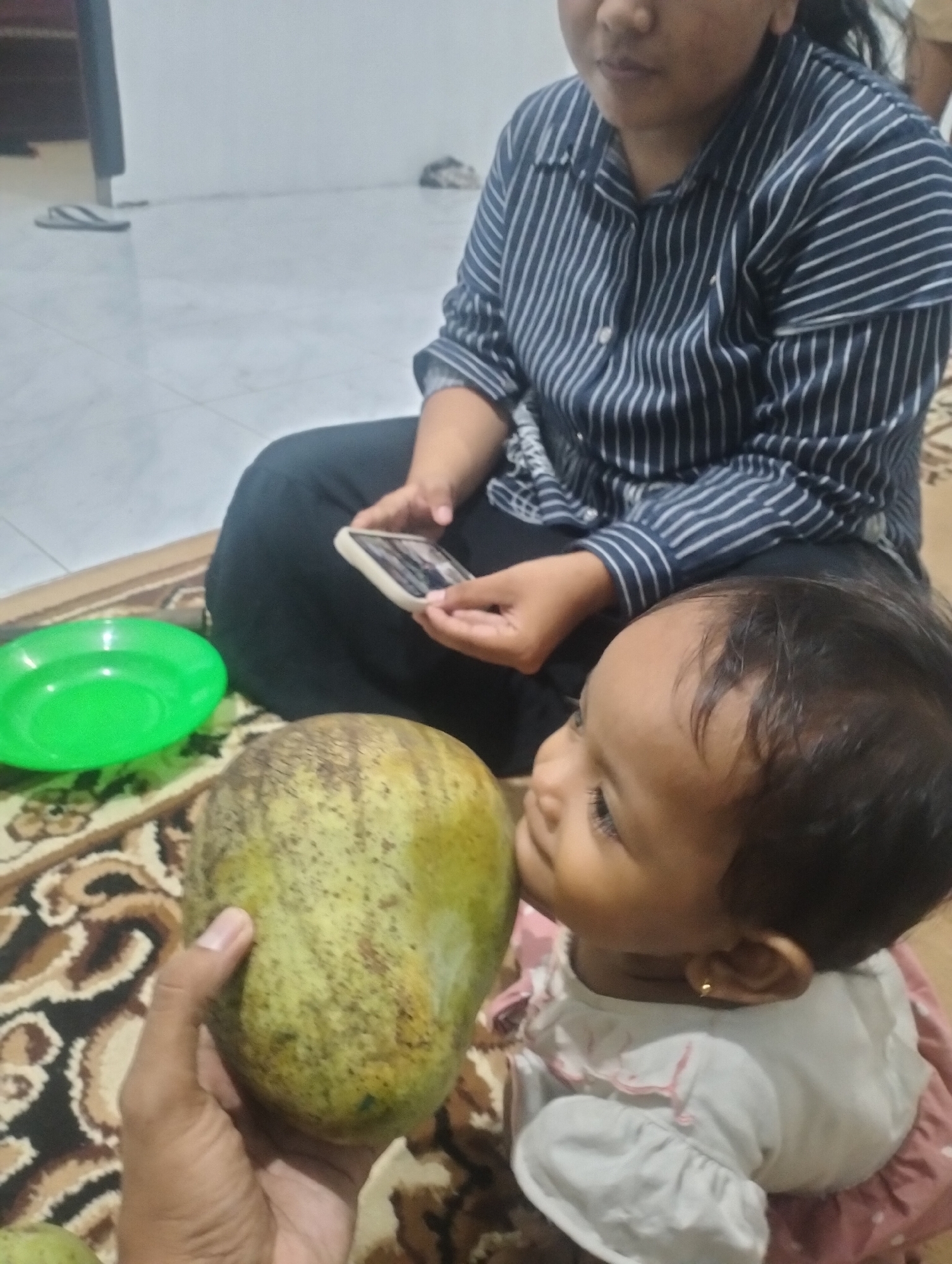 A person's hand holds a large, green and yellow mottled mango towards a young child who is looking up at it. The child is wearing a light-colored top with ruffled sleeves and a pink dress with white polka dots, and a small gold hoop earring in their left ear. Behind the child, a person wearing a dark blue and white striped long-sleeved shirt and dark pants is seated, holding a smartphone. A bright green plate is visible to the left of the mango. The scene appears to be indoors, on a patterned rug, with a white wall in the background. The location is Lhokseumawe, Indonesia.