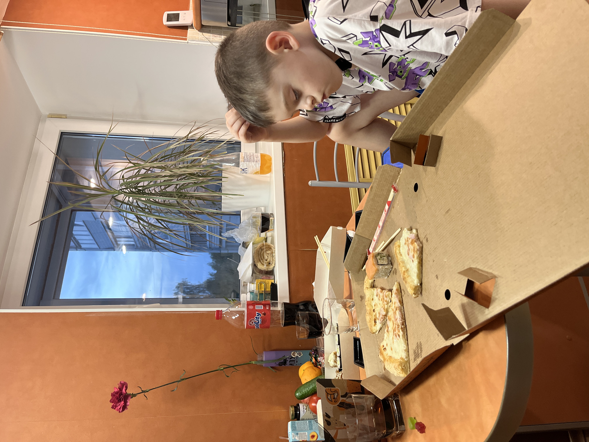 A young boy with short hair, wearing a white shirt with purple and black designs, is seated at a table in an indoor setting. He is leaning on his left arm, looking down towards a pizza box containing partially eaten pizza slices. The table is cluttered with various items including a bottle of soda, glasses, food containers, sushi, fruits (orange, cucumber), and a single red flower. A window is visible in the background, showing an exterior view with some trees and buildings. Based on the "TSARENKOV" on the boy's shirt, the location is likely in Chaikovsky, Russia.