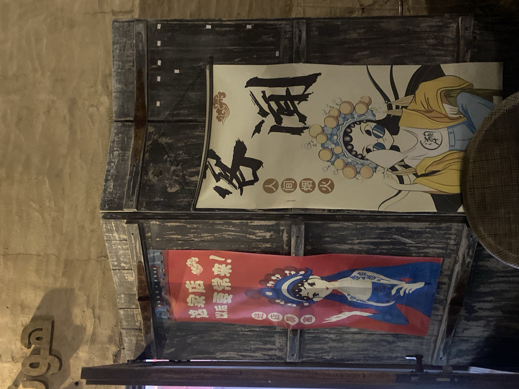 The image shows a rustic, wooden partition with two posters hanging on it. The partition appears to be constructed from aged, dark wood panels, with some signs of wear and tear. A rusty hinge is visible across the top of the partition.
On the left poster, a red background features a depiction of a woman in traditional Chinese opera attire with blue and white garments. The poster has Chinese characters written vertically in white.
On the right poster, a light-colored background displays a stylized illustration of a woman wearing a modern outfit with sunglasses and a yellow jacket. This poster also has large, bold Chinese characters in black. A small, decorative cloud motif is present in the upper right corner of this poster.
Above the partition, a sign with Arabic calligraphy is mounted on a textured, yellowish wall. The calligraphy is in a light brown color and casts a shadow.
The overall environment suggests an indoor setting with a vintage or ethnic theme, possibly a shop or restaurant. The lighting is warm and somewhat dim, contributing to the atmosphere. The location is identified as Deira, United Arab Emirates.