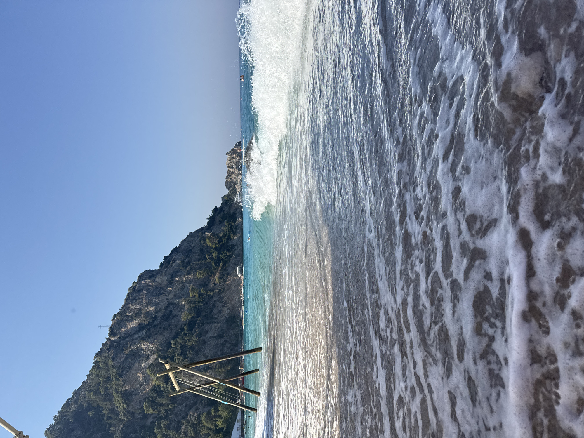 A low-angle, wide shot captures a beach scene in Fethiye, Turkey. In the foreground, gentle waves with white foam lap at a sandy shore. A large, wooden swing set stands in the shallow water to the left, its wooden posts partially submerged. In the mid-ground, turquoise water stretches towards the horizon, with a prominent wave breaking in the center of the frame, sending white spray into the air. A rocky outcrop rises from the sea in the distance. The background features a steep, tree-covered mountain slope with a clear, bright blue sky above. Several people are visible in the water, some swimming and others on small boats.