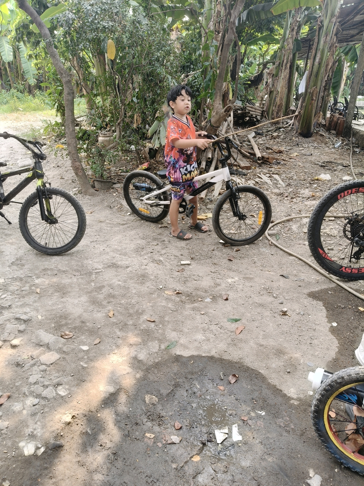 A young boy, appearing to be of Indonesian ethnicity, stands outdoors holding a stick and near two bicycles. He is wearing an orange t-shirt with a graphic print and patterned shorts. The environment is rural, with lush green foliage and trees, characteristic of the outskirts of a city like South Tangerang, Indonesia. The ground is dirt and appears to be somewhat wet in places, possibly from recent rain. There are stacks of wood and debris in the background, suggesting an area that is not meticulously maintained. The bicycles are a black and yellow BMX-style bike and a white and black bicycle. The overall impression is a casual outdoor setting where a child is playing with bicycles in a somewhat undeveloped area.