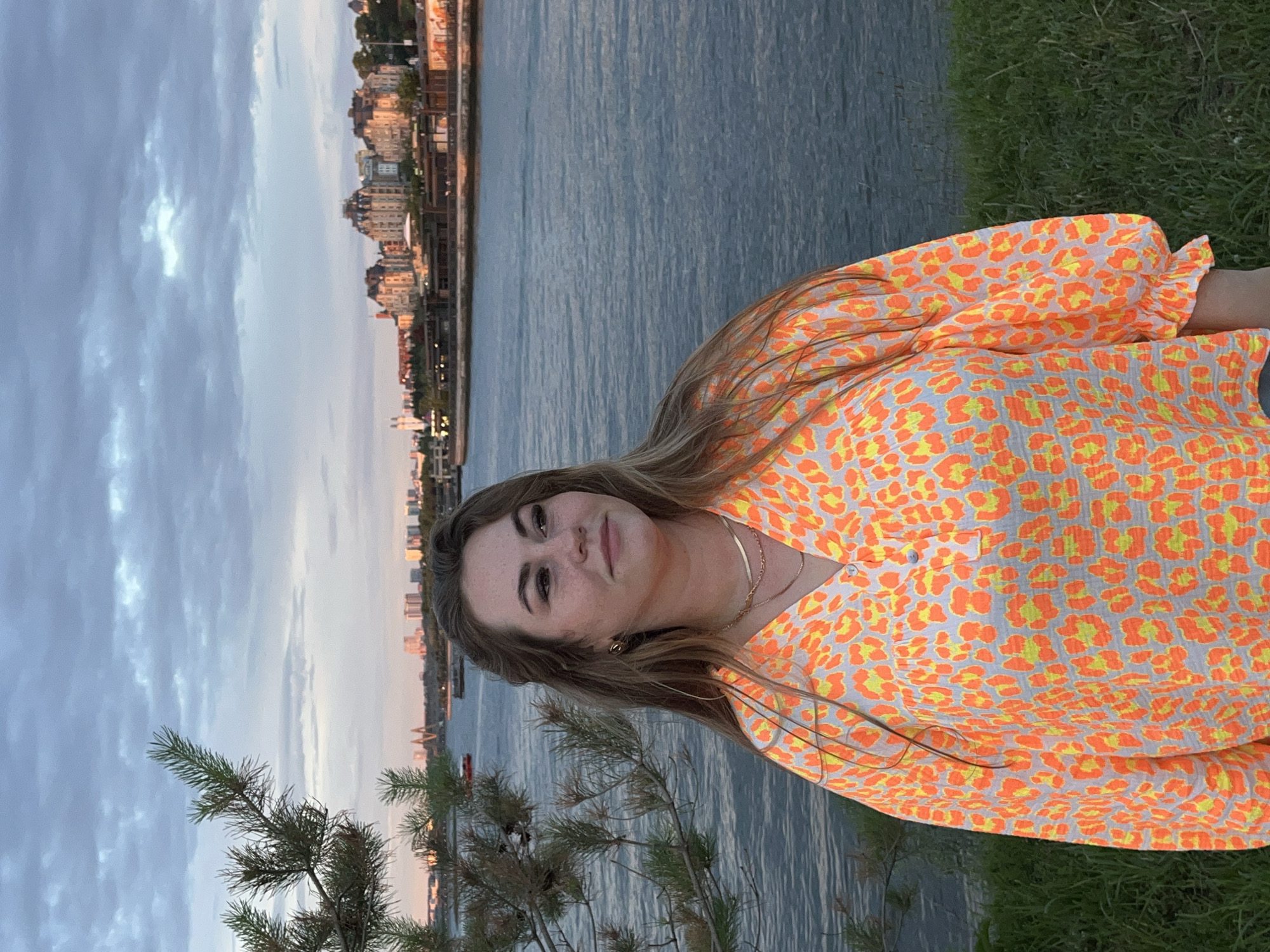 A young woman with long, light brown hair is standing in a grassy area overlooking a wide river. She is wearing a loose-fitting, long-sleeved blouse with an orange and yellow leopard print pattern. The blouse has a V-neckline and a few buttons at the top. She is wearing two delicate gold necklaces. Her left side is partially obscured by a pine tree branch.
In the background, across the river, is a cityscape with several buildings, including some with distinctive architectural styles, suggesting a European influence. The sky is cloudy with hints of pink and orange from what appears to be a sunset or sunrise. The water of the river is a deep blue-gray, reflecting the sky. The scene is calm and serene.
This location is the embankment of the Kazanka River in Kazan, Russia, specifically near the Kremlin embankment area, with the cityscape in the background likely representing the Kazan Kremlin and surrounding urban areas.