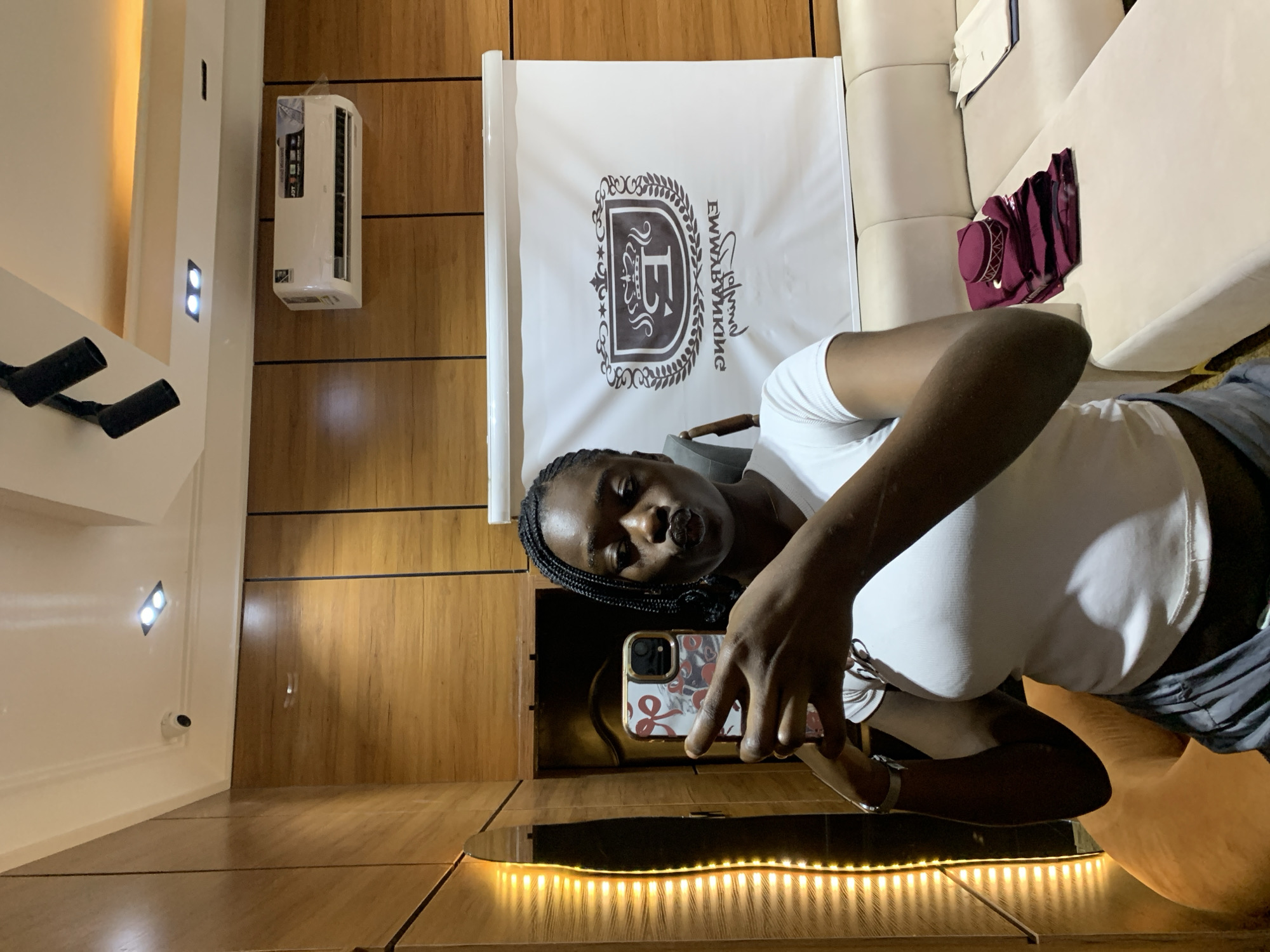 In Gura Top, Nigeria, a young Black woman with braided hair is taking a mirror selfie in what appears to be a boutique or showroom. She is wearing a white t-shirt and grey pants, and is puckering her lips as she holds up her phone, which has a patterned case. To her left, a wall is illuminated by vertical LED strip lights. Behind her, a large white banner with a crest and text, likely a logo, is displayed against a wood-paneled wall. An air conditioning unit is visible on the upper part of the wall. A light-colored sectional sofa is in the foreground, with a stack of folded maroon fabric and a maroon cap, as well as a beige shirt, resting on it.
