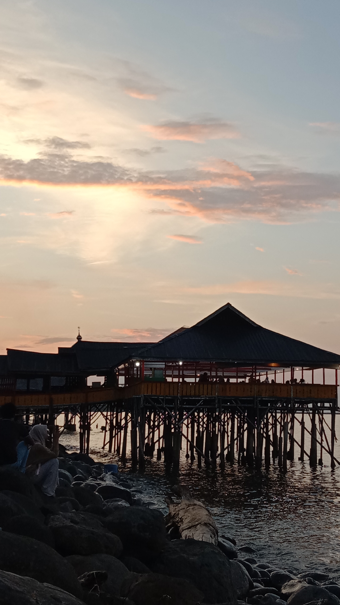 An atmospheric wide shot captures a serene coastal scene in Lhokseumawe, Indonesia, at twilight, likely sunset. The sky is painted with soft pastel hues of light blue, pink, orange, and yellow, adorned with scattered, wispy clouds.
The main subject is a large stilt structure, likely a restaurant or café, with a dark, peaked roof, extending over calm waters. It is supported by numerous dark wooden stilts. The interior of the building is softly illuminated, revealing the silhouettes of several people, presumably patrons, enjoying the establishment's ambiance.
In the immediate foreground, a rocky shoreline borders the tranquil water. A person, appearing to be a woman wearing a light-colored head covering (hijab), a brown top, and patterned trousers, sits on the dark rocks to the left. She seems to be relaxing and observing the view, with a light blue backpack resting beside her. A piece of driftwood or a fallen tree trunk is also visible among the rocks near the water's edge.
The scene conveys a peaceful and contemplative mood as day transitions to night, highlighting leisure activities by the sea. There is no visible text in the image.
