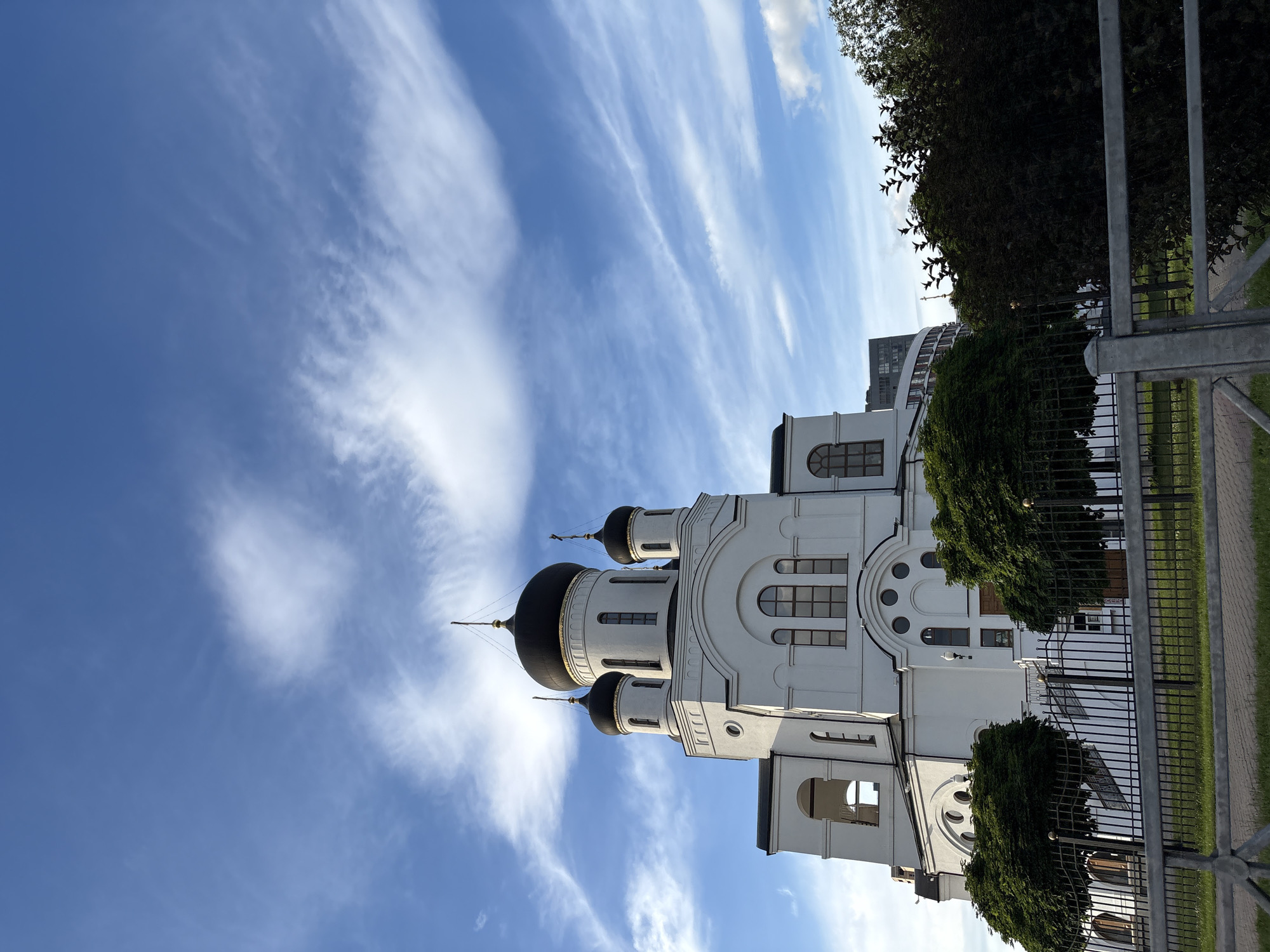 The image depicts a white Orthodox church with black domes, set against a bright blue sky filled with wispy white clouds. The church architecture is characterized by arched windows and multiple cupolas. The scene is bordered by a metal fence and lush green trees. To the right of the church, a modern, curved building is visible in the background. The lighting suggests it is daytime, with the sun casting shadows on the building and illuminating the sky. The overall atmosphere is serene and tranquil. No people are visible in the image, and no text is present. The orientation of the image is vertical, with the sky occupying the upper portion and the church and surrounding elements filling the lower part. The setting appears to be a public space, possibly a park or a church grounds, in Chelyuskinsky, Russia.