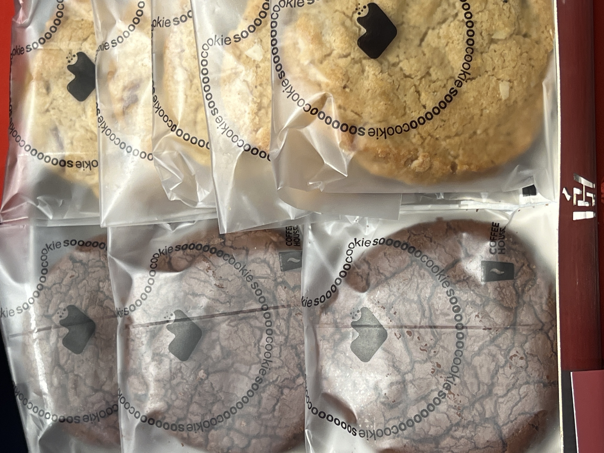 This image shows a box of individually packaged cookies. The cookies are in clear plastic wrappers with the logo "cookie soooooo" printed in a circular pattern around the cookie. There are two types of cookies visible: lighter, beige-colored cookies and darker, chocolate-colored cookies with a cracked surface. The cookies appear to be inside a red box. The logo "COFFEE HOUSE" is visible on one of the darker cookies, suggesting a possible association with a coffee shop. The city of Yerevan, Armenia, cannot be confirmed from this image alone.