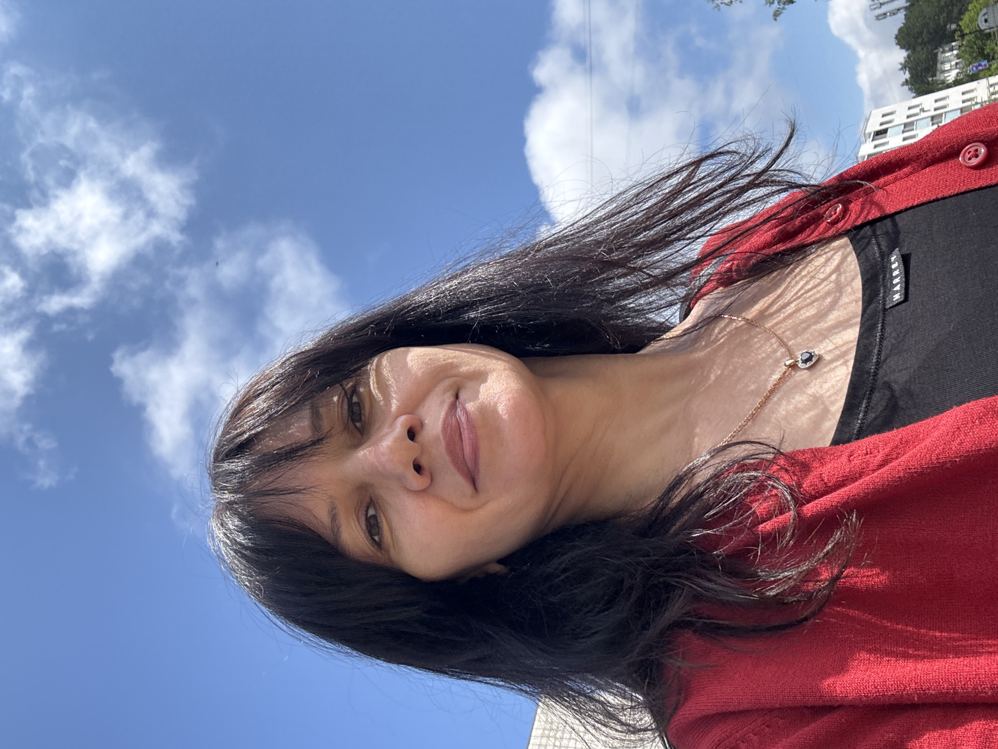 The image displays a woman photographed outdoors on a bright, sunny day, captured from a low angle. She has long, dark hair with bangs, dark eyes, and a pleasant, subtle smile, with her head slightly tilted. Her hair appears to be gently blowing, suggesting a light breeze. She is wearing a red cardigan with visible red buttons over a black top. A tag on the black top's left shoulder is visible, bearing the text "MARREXX". Around her neck is a delicate gold chain with a small, dark-stoned pendant.
The background features a clear blue sky with scattered white clouds. Distant architectural elements, including a multi-story white building with visible windows, and some trees are faintly visible on the right side of the frame. The strong sunlight indicates it is daytime. The scene suggests an outdoor setting in an urban or suburban environment, consistent with a location like Chaikovsky, Russia.