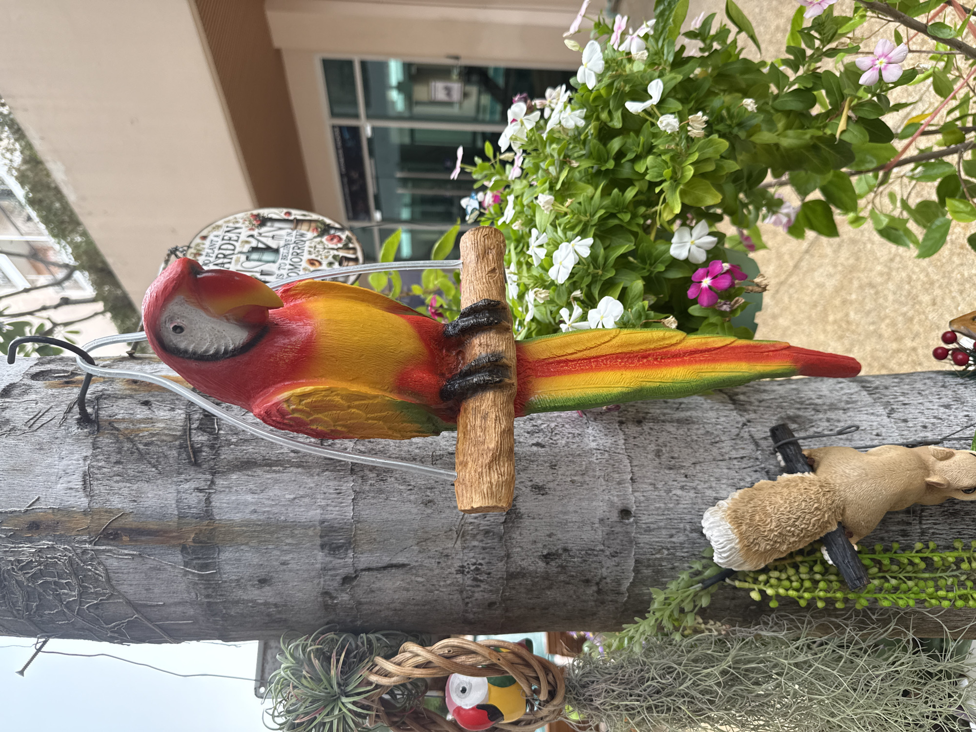 The image captures a decorative ceramic parrot, brightly colored in red, yellow, and green, perched on a wooden branch attached to a palm tree. The parrot is hanging from a wire. To the left of the parrot, a small, decorative birdhouse with a ceramic bird inside is suspended from the tree. Below this, several air plants hang, some with stringy, grey foliage. A small, brown ceramic squirrel is clinging to the trunk of the palm tree below the parrot's perch, holding a dark branch. In the background, a planter filled with lush green foliage and blooming white and pink flowers is visible. Further in the background, a building with glass doors and windows is partially visible, suggesting an outdoor commercial or residential area. A circular garden sign with text that reads "TO PLANT A GARDEN IS TO BELIEVE IN TOMORROW" is hanging behind the parrot. The overall scene appears to be an outdoor decoration in a tropical or subtropical setting, consistent with Pattaya City, Thailand. There are no people visible in the image.