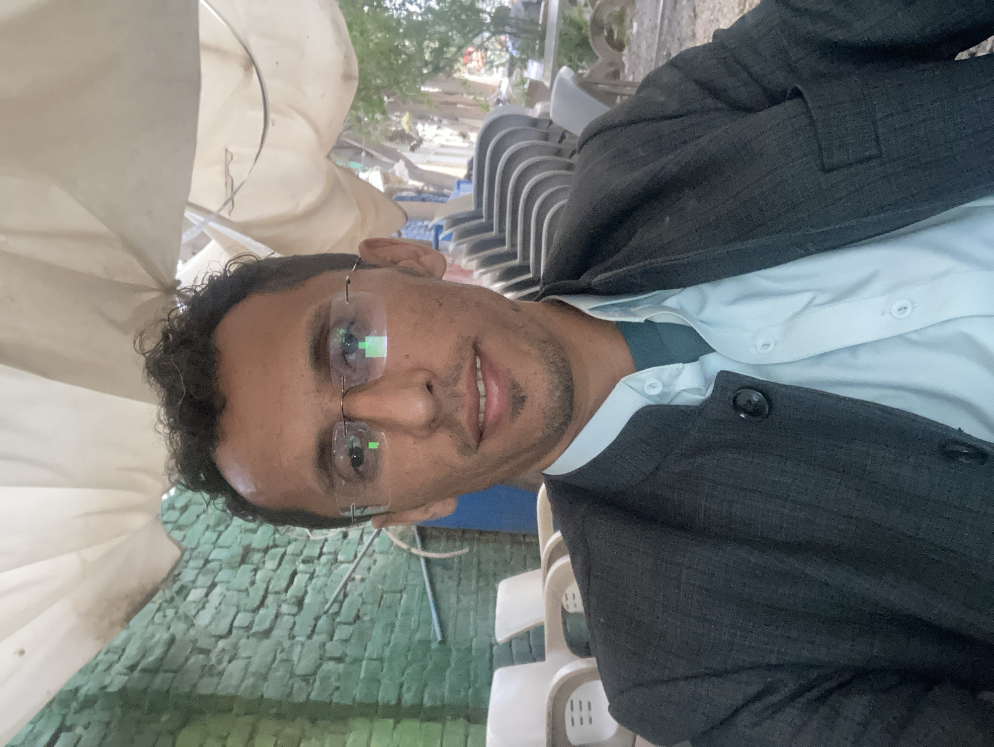 The image depicts a male individual, appearing to be in Quetta City Tehsil, Pakistan, in a semi-outdoor setting. He is positioned centrally, facing the camera directly with a faint smile. The subject has dark, curly hair and wears clear-framed glasses, which exhibit distinct green and white reflections. His attire consists of a light blue button-up shirt, open at the collar, layered over a darker undershirt, and a dark grey, textured jacket with visible black buttons on the front closure. The background on the left features a teal-painted brick wall against which two light-colored plastic chairs are visible. To the right, a stack of approximately five to seven light grey plastic chairs is present, with blurred foliage, trees, and a dirt ground visible beyond. An off-white fabric canopy or awning provides overhead cover. No other individuals are present.
