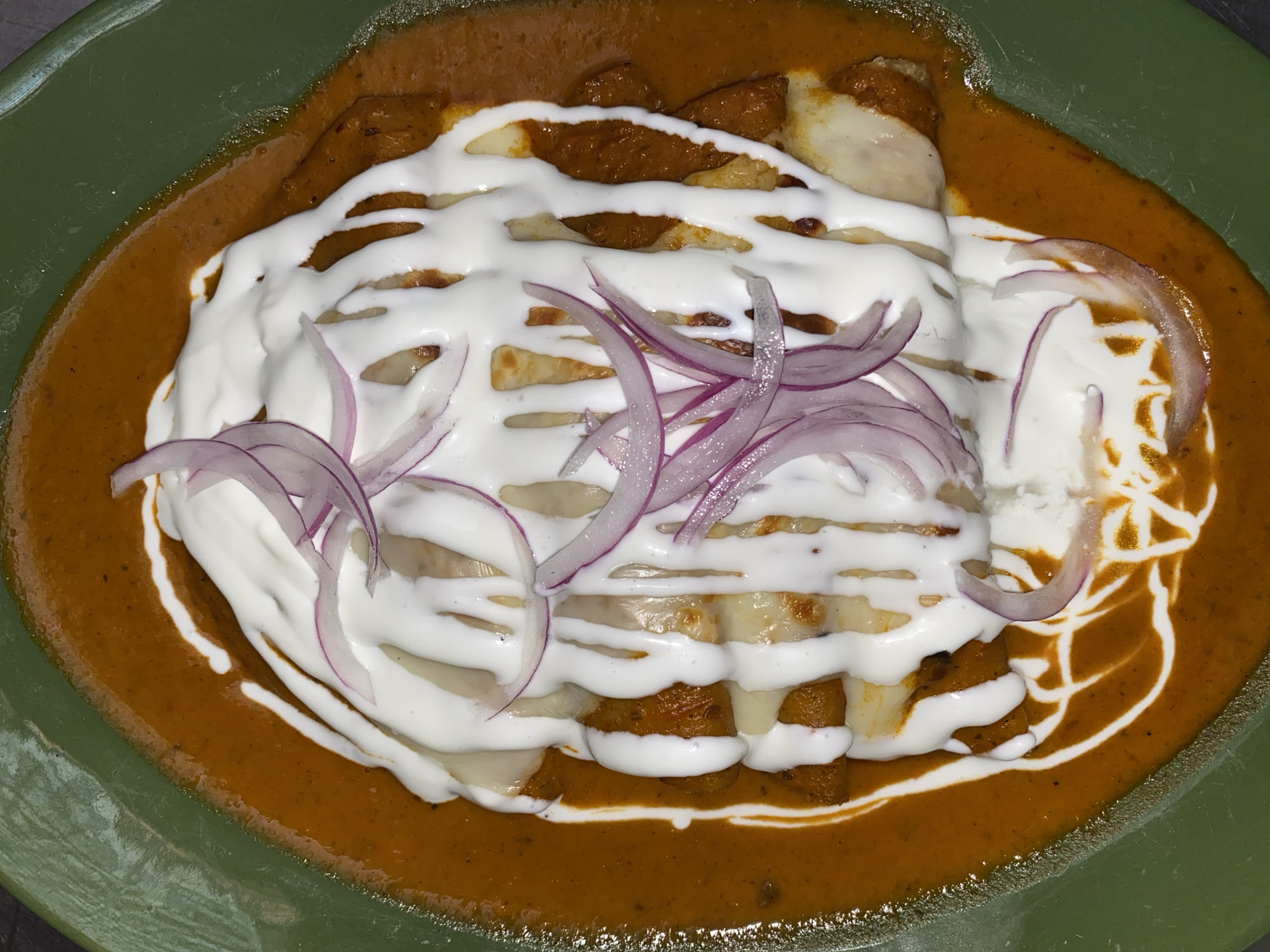 An overhead shot depicts a green plate of enchiladas poblano, potentially located in the city of Puebla, Mexico. The enchiladas are bathed in a brown sauce, likely mole poblano, and drizzled with white crema or sour cream, arranged in parallel lines. Thin slices of red onion garnish the top of the enchiladas. Cheese is visible between the enchiladas and the crema.