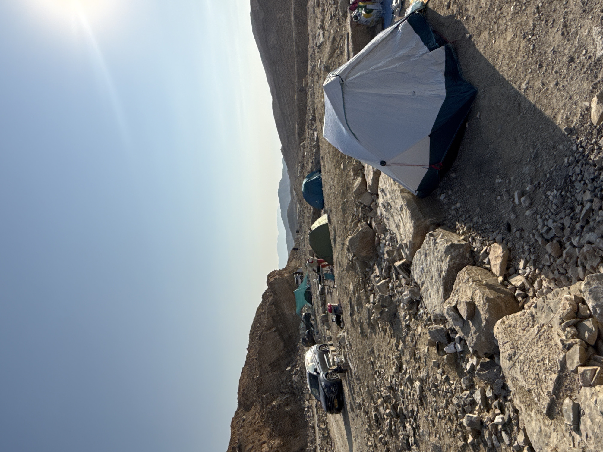 This image depicts a desert campsite in the Ramat Negev Regional Council, Israel. Several tents are set up on rocky, arid terrain. Two dark-colored SUVs are parked near a cluster of tents and a picnic table, suggesting an outdoor expedition or camping trip. The sun is visible in the upper right corner, casting long shadows across the scene. The landscape is characterized by barren hills and mountains, typical of the Negev Desert. The sky is clear and blue, indicating fair weather conditions.