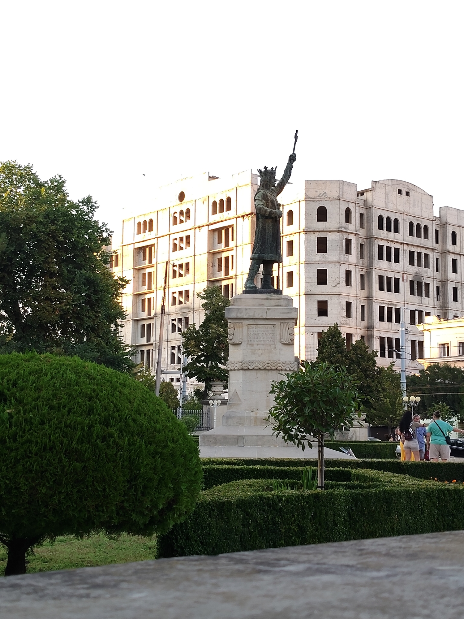 The image shows a statue of Stephen the Great in Chișinău, Moldova. The statue is located in front of a building that is under construction. The building has a neoclassical style with arched windows and columns. There are trees and bushes around the statue, and a group of people is standing in the background. The sky is clear and bright.