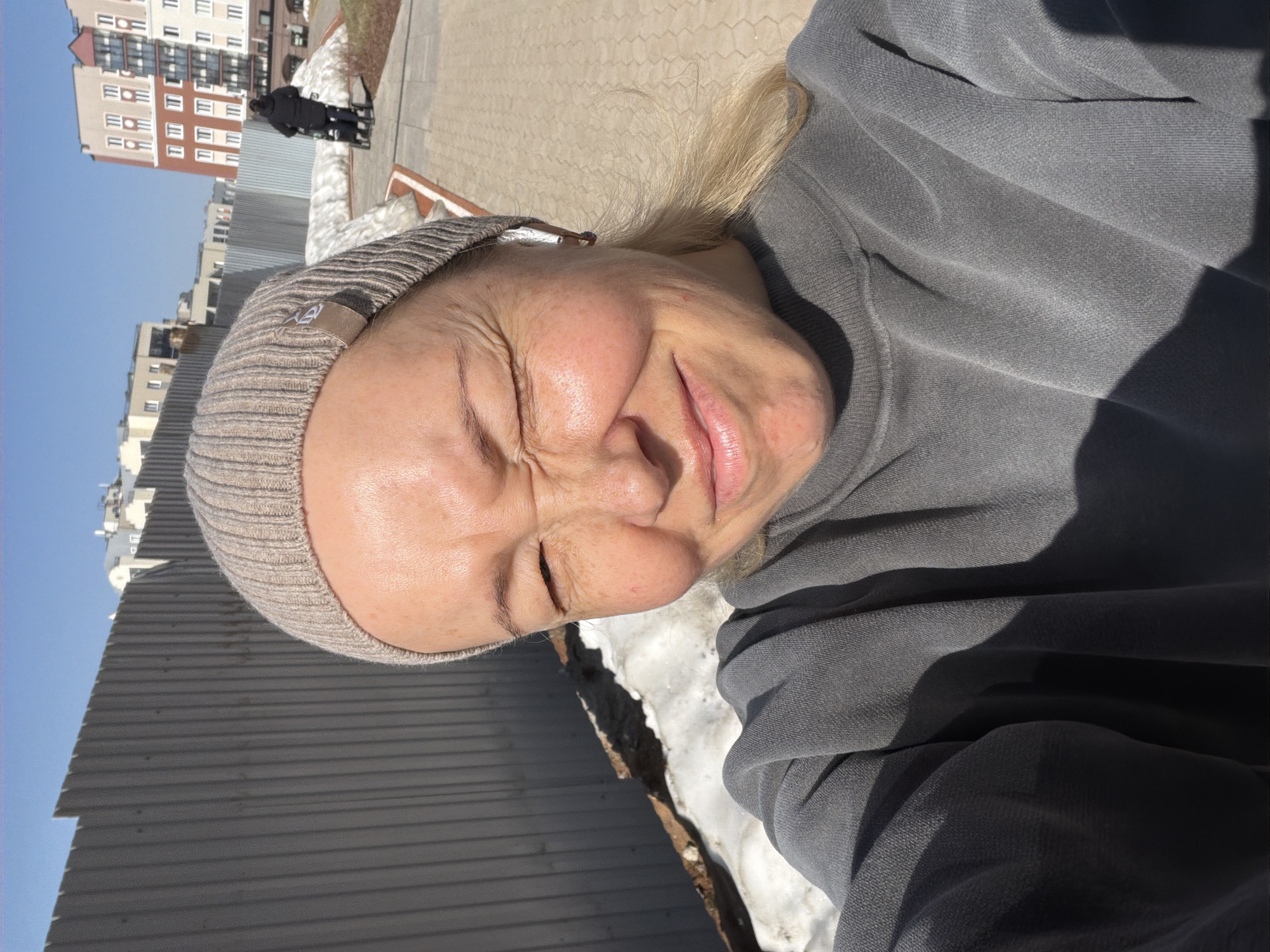 A selfie photograph shows a middle-aged woman wearing a knitted beanie and a gray sweatshirt. The background suggests an urban environment in Pervomaysky, Russia, with buildings visible in the distance. There is melting snow in the foreground, and a corrugated metal structure is also visible. The woman's eyes are closed and her face is partially obscured by shadow, possibly due to the bright sunlight.