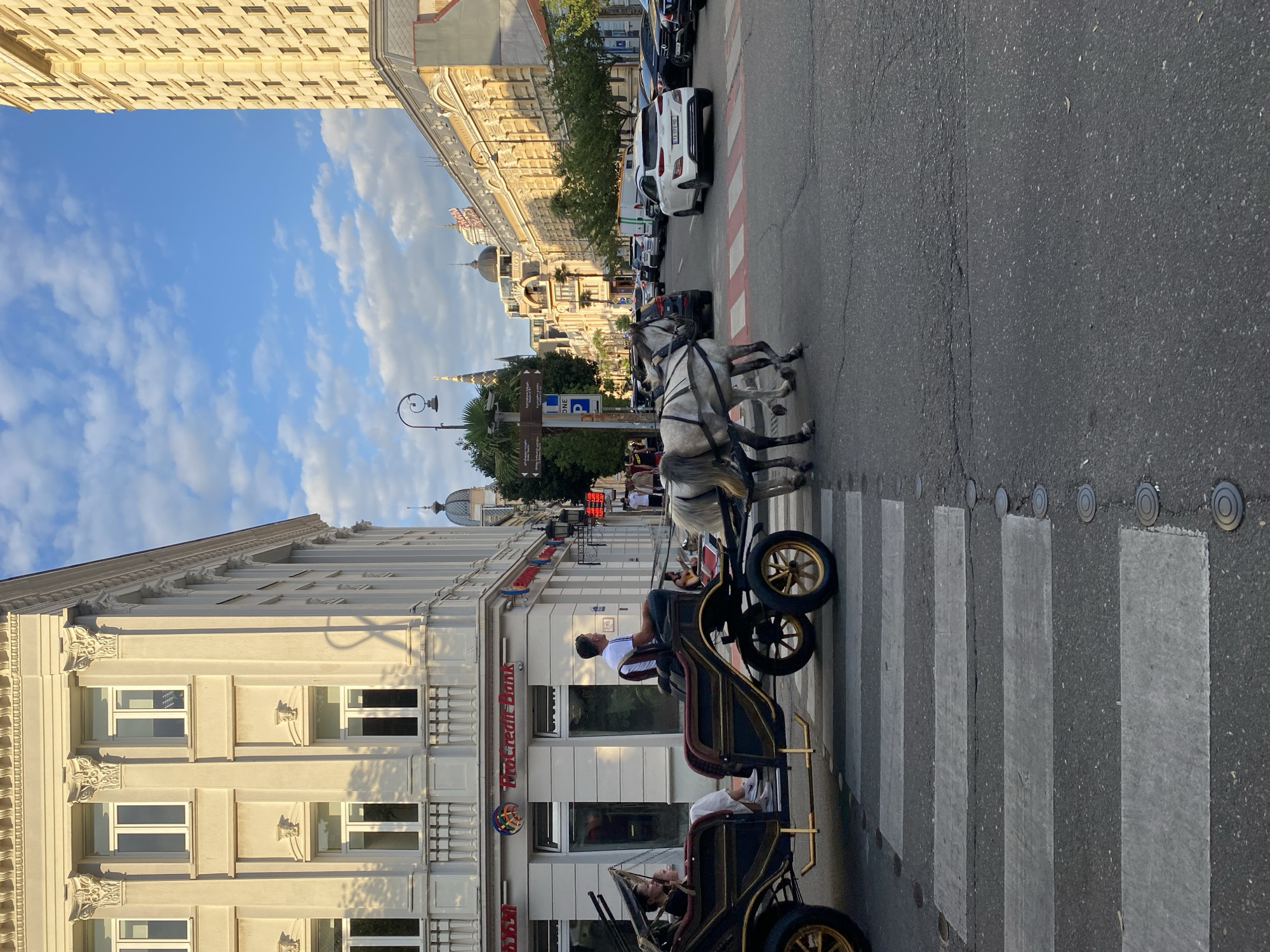 The image depicts a street scene in Batumi, Georgia, likely near the intersection of Rustaveli Avenue and Ninoshvili Street, given the architecture and the presence of horse-drawn carriages. In the foreground, a horse-drawn carriage with two passengers, a woman and a child, is crossing a pedestrian walkway. The carriage is pulled by two grey horses. The buildings lining the street are a mix of classical and modern architectural styles, with the building on the left featuring columns and ornate detailing, and the building on the right being a tall, modern structure. Several cars are also visible on the street, indicating it is a busy urban area. The sky is blue with scattered clouds, suggesting it is daytime. The overall impression is of a tourist-friendly area in Batumi, where traditional horse-drawn carriage rides are offered to visitors.