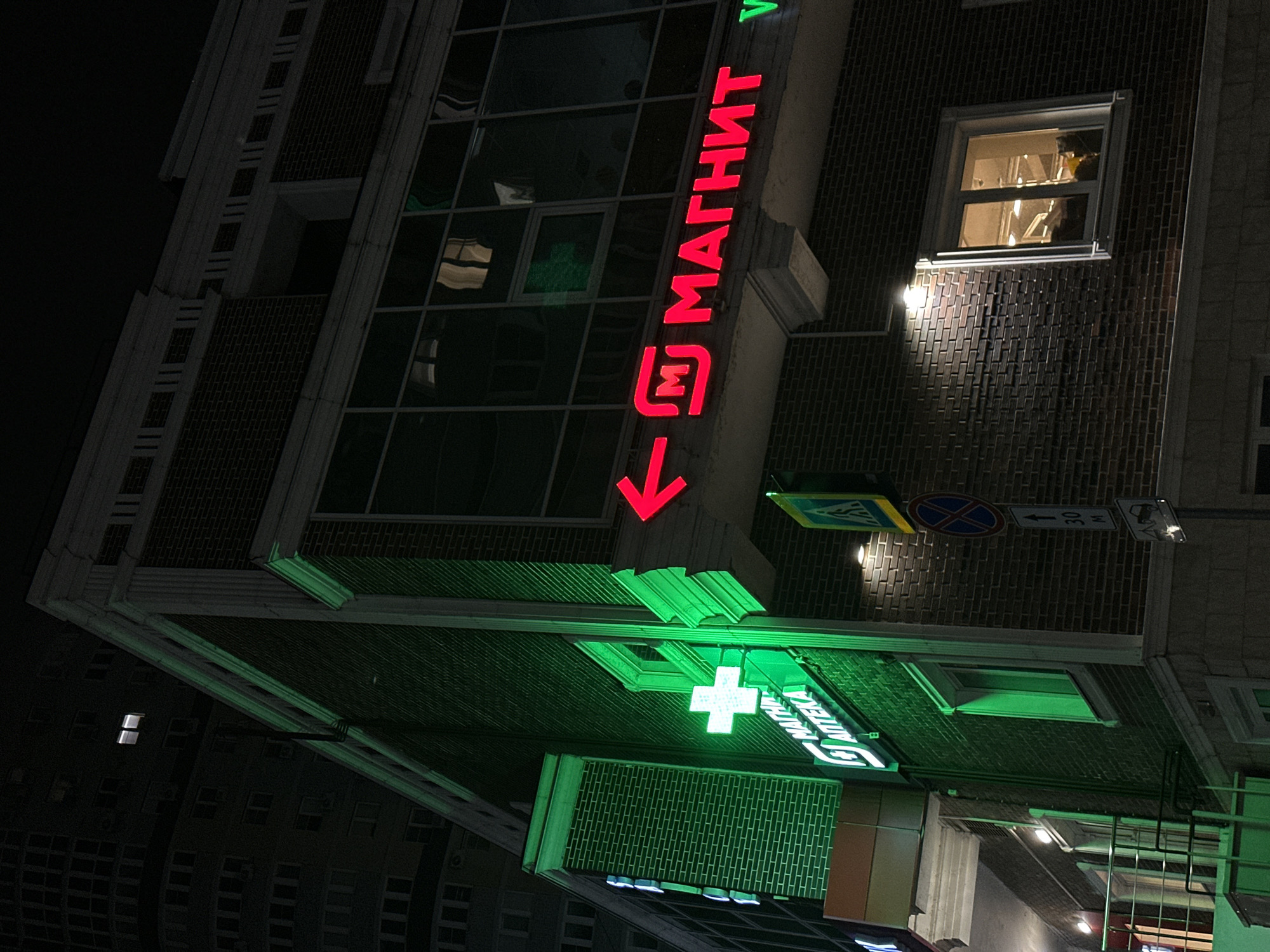 A low-angle, tilted, long-exposure photograph depicts the illuminated facade of a building at night in Rostov-on-Don, Russia. The building appears to be a commercial structure, possibly a multi-use building housing retail and other businesses. The red, vertically stacked neon sign "МАГНИТ" (Magnit - a Russian retail chain) with an arrow points downwards to a green-lit pharmacy (APTEKA) sign with a medical cross. Traffic signs are above. The architecture of the building includes dark brickwork. Window shows activity inside the building. Other buildings are visible in the background.