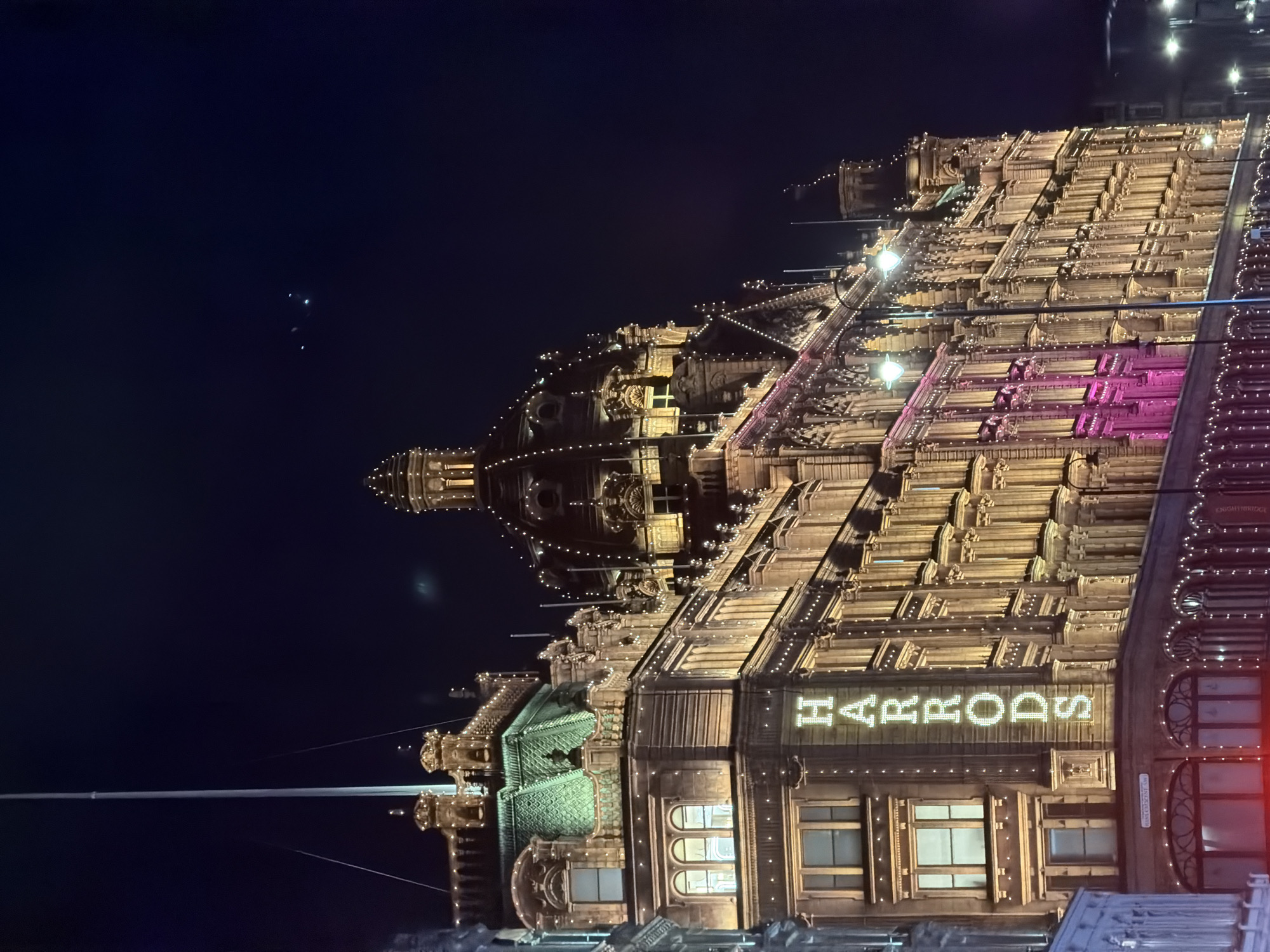 The image depicts Harrods department store in Knightsbridge, London, at night. The building's facade is elaborately illuminated with lights, and the name "Harrods" is prominently displayed in illuminated lettering. The architectural details of the building are visible, including windows, moldings, and decorative elements. The sky is dark.