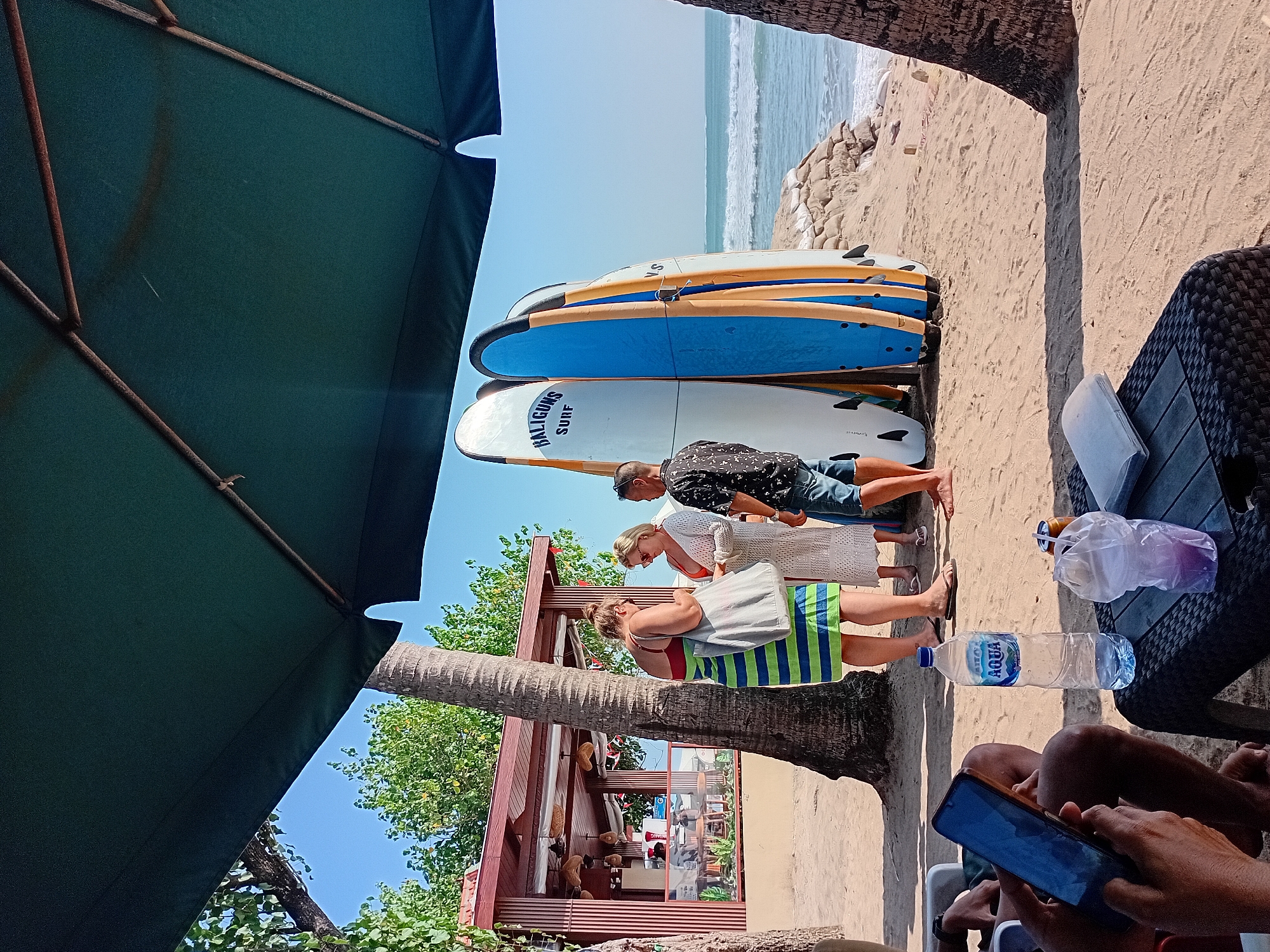 This image depicts a beach scene in Kuta, Indonesia. In the foreground, a person's hands are visible holding a smartphone. A black woven table is situated in front of them, holding a bottle of Aqua water, a drink in a plastic cup with a straw, and a slim metal can. Beyond the table, four surfboards are propped up vertically on the sand. The surfboards are white, blue, and yellow. Behind the surfboards, a group of three people, two women and one man, are conversing. The woman on the left wears a green and blue striped towel around her waist and a red bikini top, carrying a gray tote bag. The woman next to her is dressed in a white lacy cover-up and sunglasses, and the man is wearing a black patterned shirt and denim shorts. To the left of the people, a thatched-roof building with open sides, likely a beach bar or cafe, is visible, with palm trees in the background. The ocean and waves can be seen in the distance, under a clear blue sky, with the edge of a large green umbrella partially obscuring the top of the frame. The scene is set on a sandy beach.