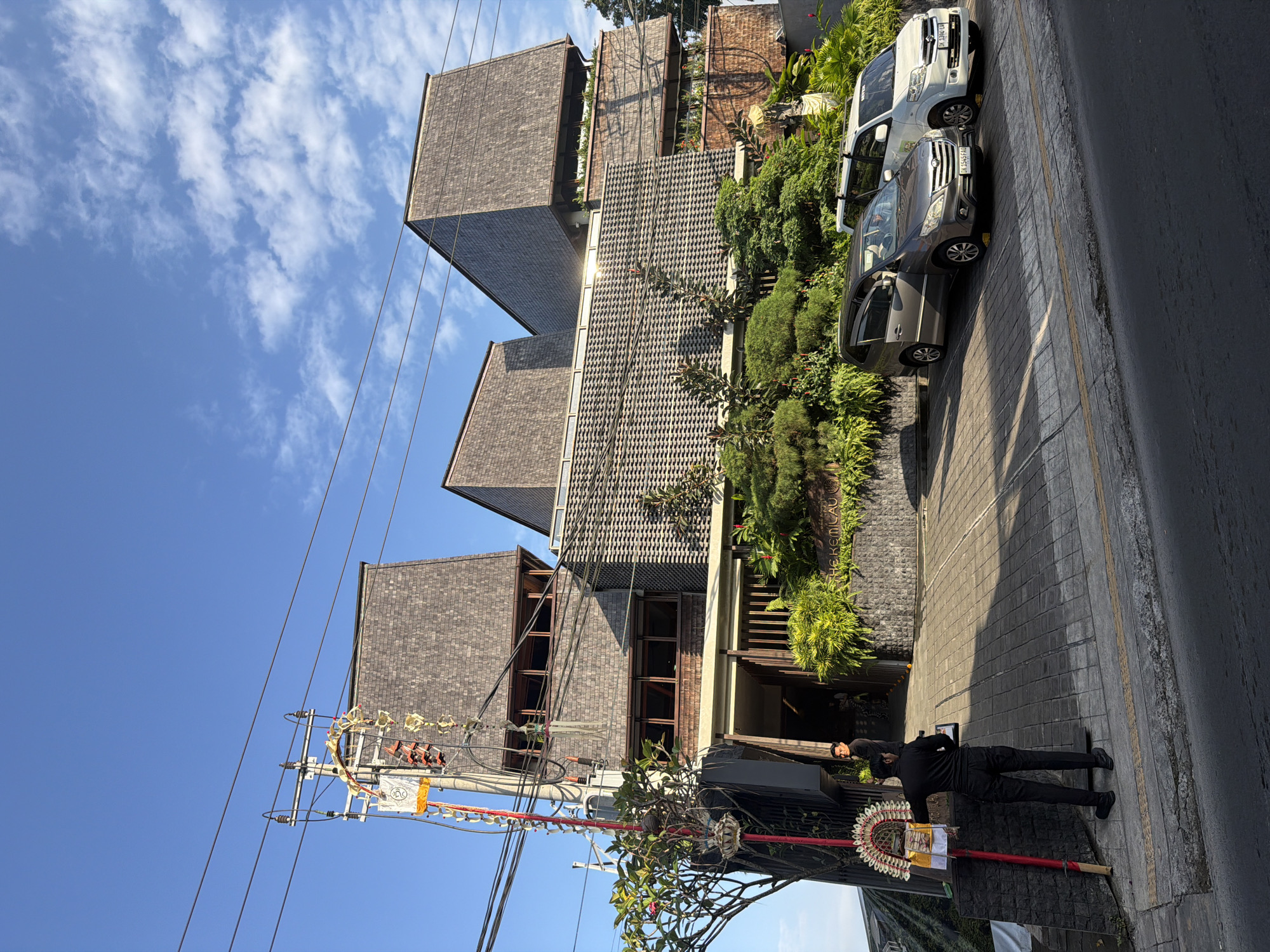 The image depicts the exterior of a multi-story building with a distinctive architectural style, featuring angled roofs covered in dark shingles and textured grey facade elements. The building appears to be a commercial establishment, possibly a hotel or restaurant, as suggested by the sign "THE KEMILAU" visible near the entrance. In front of the building, a paved driveway leads to the entrance, with several cars parked on the side. A man in dark clothing stands near the driveway, holding a clipboard, possibly an employee. Another man in similar attire is visible near the entrance of the building. The scene is illuminated by natural sunlight, casting shadows and highlighting the textures of the building and surrounding vegetation. Lush green plants and trees are incorporated into the building's design, creating a natural aesthetic. The overall impression is a modern, stylish building set against a clear blue sky with scattered clouds, indicative of a location in a tropical climate. The presence of a utility pole with overhead wires and decorative elements suggests an urban or semi-urban setting. The scene is likely located in Ubud District, Indonesia, a region known for its cultural attractions and hospitality businesses.