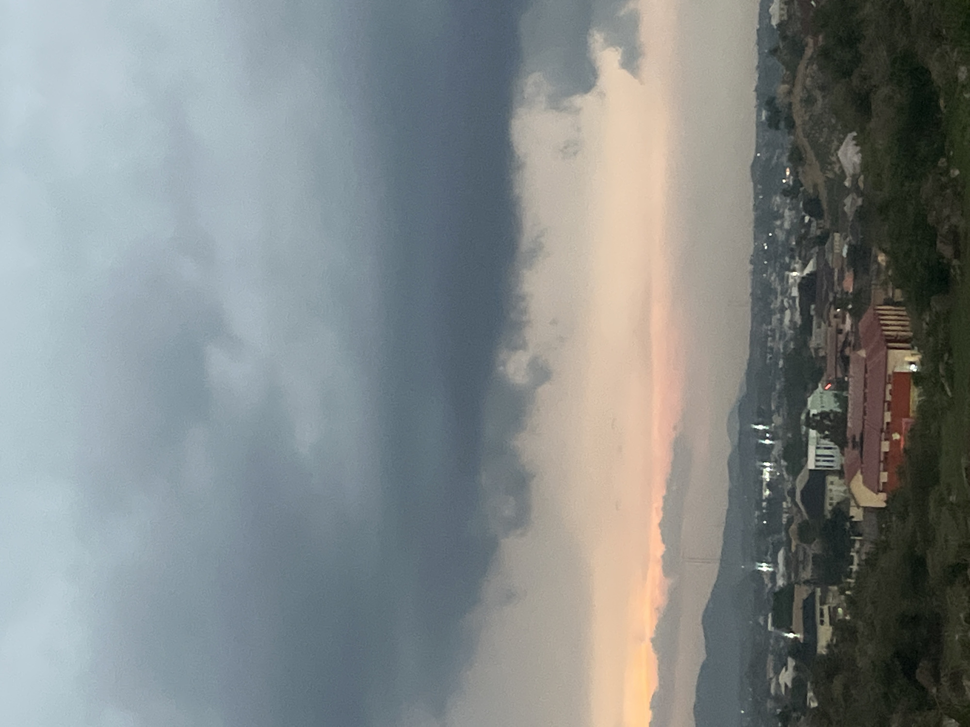 The image shows a panoramic view of the city of Gura Top, Nigeria, at dusk. Dark, heavy clouds fill the upper portion of the sky, with a gradient of lighter hues near the horizon, suggesting the setting sun. Below the clouds, a city sprawls across rolling hills. Numerous buildings with red and white roofs are visible, interspersed with darker structures and greenery. Some buildings have illuminated windows, indicating activity within. The landscape is dotted with trees and shrubs, especially in the foreground, which is rocky and uneven. In the distance, mountains form a hazy backdrop against the fading light. The overall scene depicts a calm urban environment as evening descends.