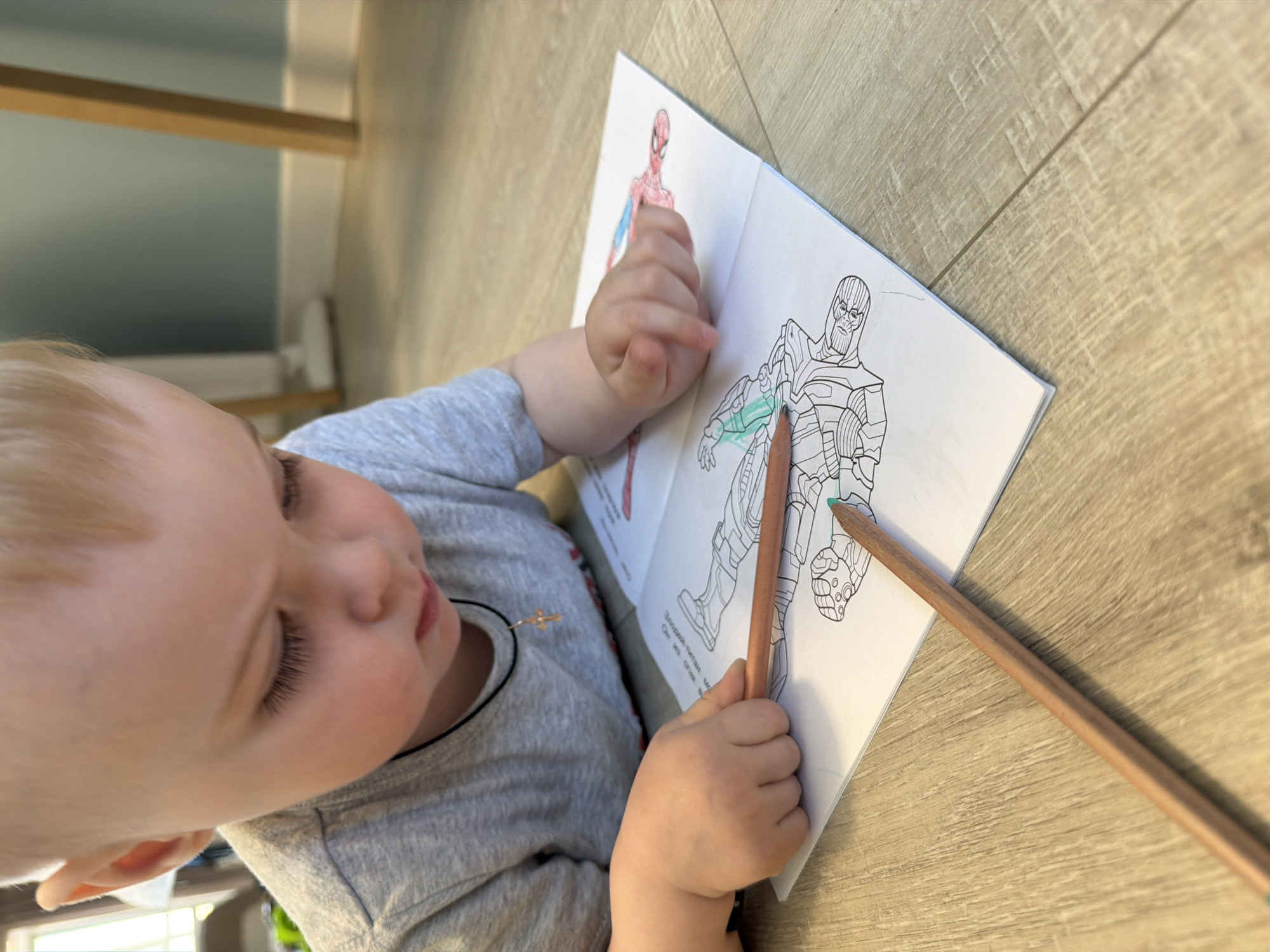A young child with blond hair is coloring in a book. The child is wearing a gray t-shirt and a necklace with a cross. The book is open to a page with a drawing of Spider-Man on the left and a drawing of Thanos on the right. The child is using a green pencil to color Thanos. There is another pencil lying on the table next to the book. The background is a light brown wood-grain surface. The location is unknown.