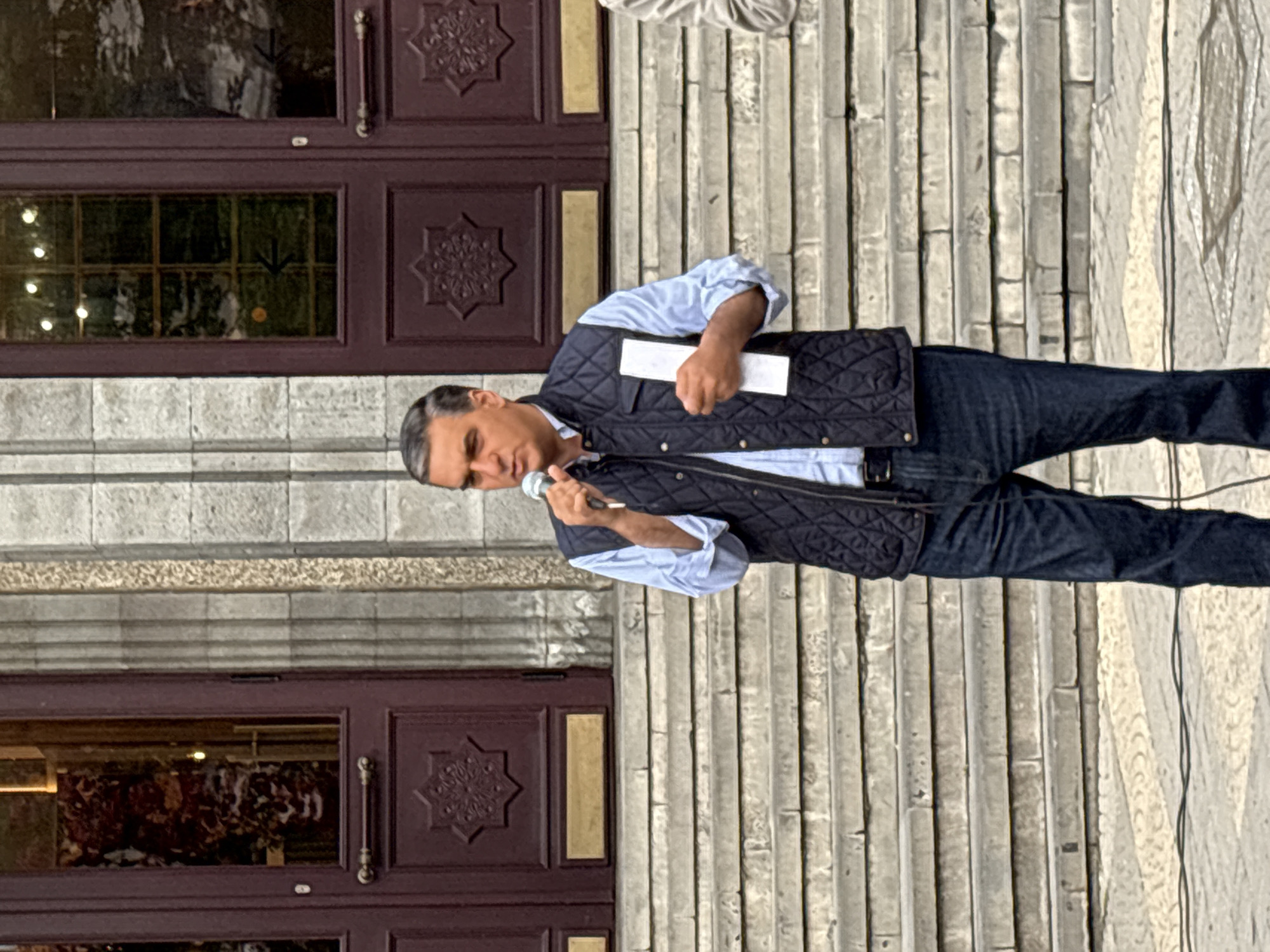 A man, seemingly in his 50s or 60s, is speaking into a microphone while standing in front of a building. He is wearing a light blue shirt, a dark quilted vest, and dark jeans. In his left hand, he holds a white piece of paper or a card. The building behind him is a light-colored stone structure with large, ornate doors, possibly a government building or a cultural center. The architecture indicates a location in Yerevan, Armenia, based on the typical Soviet-era construction style and design elements often found in the city. The man appears to be giving a speech or making an announcement outdoors.