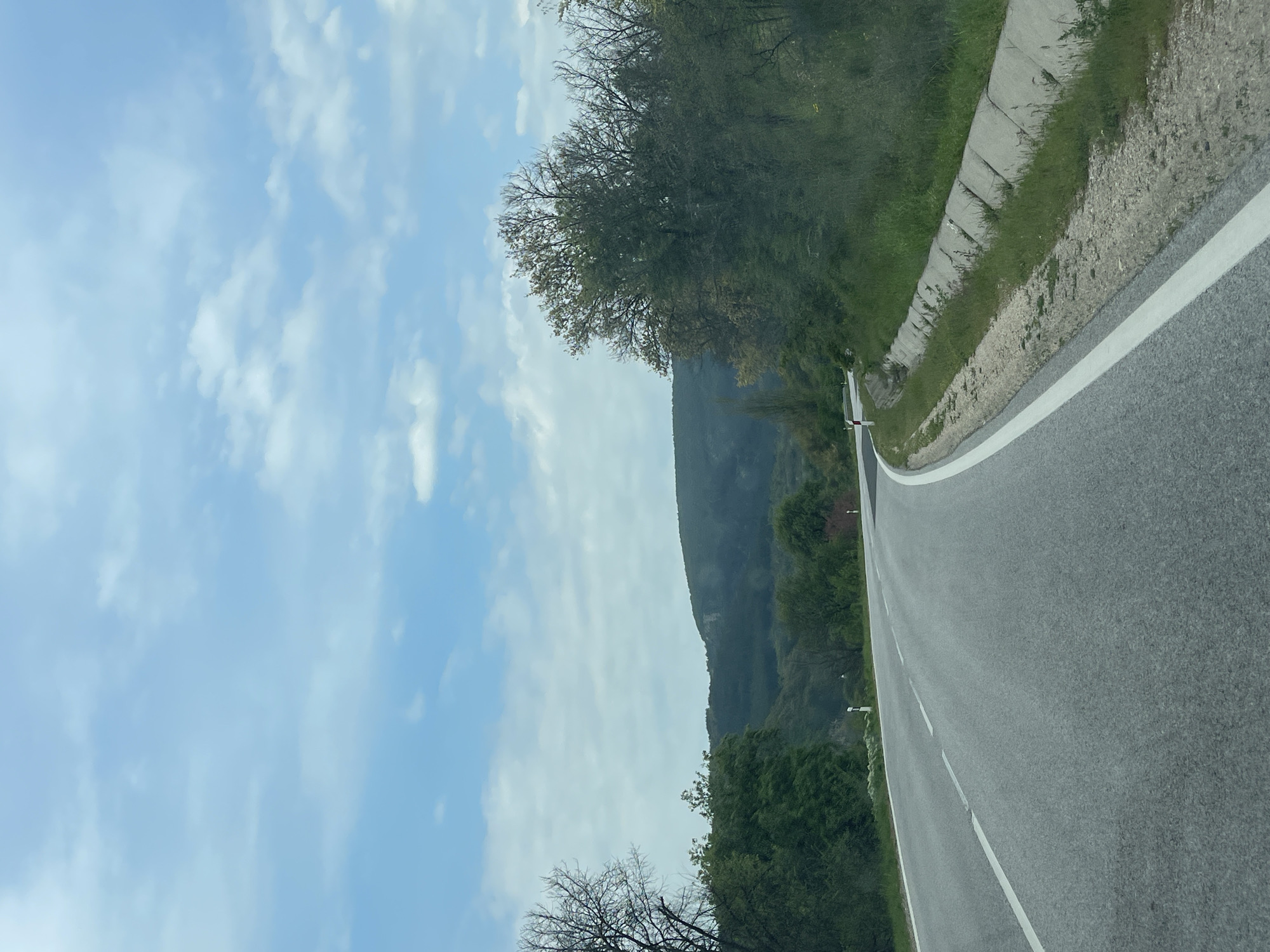 This image, captured in Madrets, Bulgaria, depicts a serene, winding asphalt road traversing a natural, mountainous landscape. The perspective suggests it was taken from a moving vehicle, looking ahead along the road.
In the foreground and mid-ground, the road features a clear solid white line marking its right edge and a dashed white line, possibly separating lanes or indicating a shoulder. To the immediate right of the road, a concrete curb or drainage channel runs alongside, bordered by green grass and light-colored earth. Lush green vegetation and a variety of trees line both sides of the road. Some trees exhibit sparse, dark branches, indicating either early spring or deciduous species, while others are full of vibrant green foliage.
In the background, a large, dark green mountain or hill rises majestically against the sky, its slopes densely covered in trees. The sky above is a bright, clear blue, adorned with scattered, fluffy white clouds, suggesting fair weather conditions. The natural lighting indicates it is daytime, likely morning or afternoon, given the soft, even illumination.
No people or visible text are present in the image, emphasizing the tranquil and undeveloped nature of the location. The scene evokes a sense of peaceful travel through a picturesque, rural or mountainous environment.
