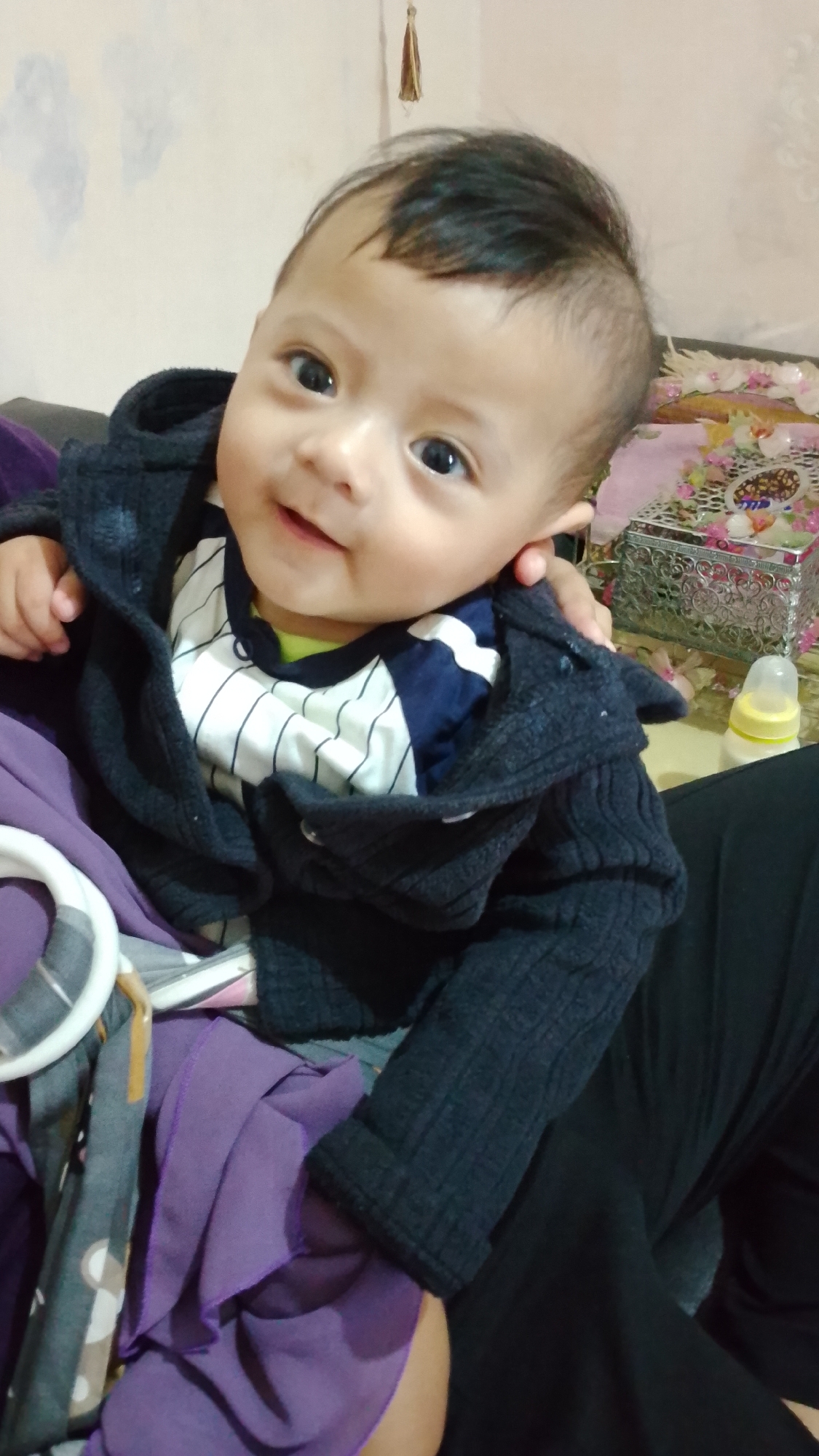 A young child is seated, wearing a dark blue hooded jacket over a striped shirt, and is wrapped in a purple cloth. The child is looking towards the camera with dark eyes and a slight smile. The background is blurred but appears to be an indoor setting, possibly a living room, with furniture and decorative items visible. A baby bottle is on a table in the background. The image was taken in Bojongsoang, Indonesia.