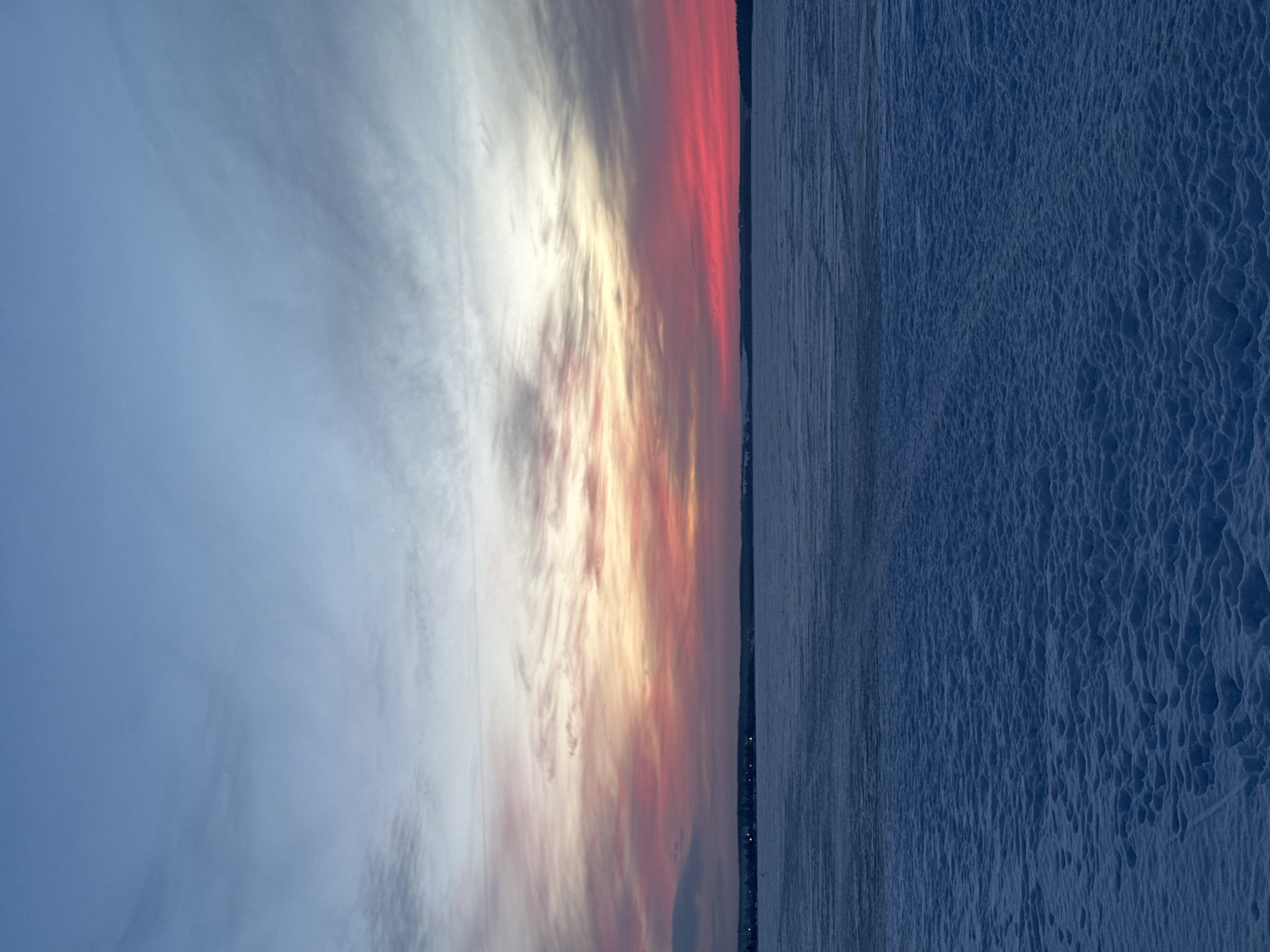 The scene captures a wide exterior view of a winter twilight over the Votkinsk Reservoir in Votkinsk, Russia. The lower half of the frame presents a dark blue ground plane covered in snow, exhibiting an uneven, undulating texture with irregular depressions. Above this, a flat, lighter blue expanse, identified as frozen water, extends to the horizon. A narrow, dark band of land, likely the distant shoreline, is visible at the horizon, featuring several faint points of artificial illumination. The upper portion of the image is dominated by the sky, displaying a stratified coloration indicative of twilight. Directly above the horizon, a band of deep red and magenta hues transitions upward through orange and yellow tones, fading into pale grey and light blue at the zenith. Wispy, cirriform clouds are present across the sky, reflecting these atmospheric colors. No human figures, vehicles, or distinct man-made objects are visible within the immediate foreground or midground.