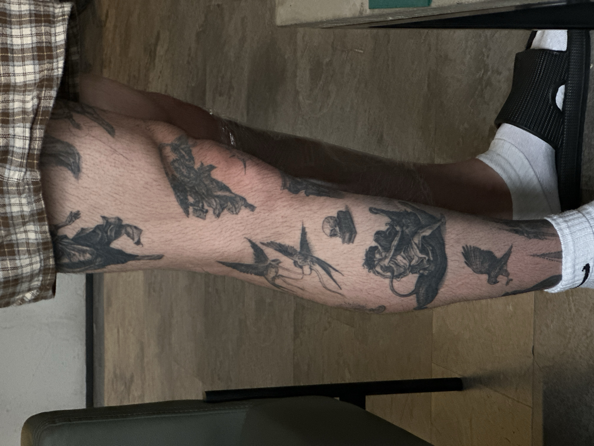 The image shows a person's leg covered in tattoos, likely sitting down. The leg has several visible tattoos depicting birds and other graphic designs. The person is wearing a white sock and a black sandal. The environment appears to be indoors with light brown walls and flooring. The presence or location in Moscow, Russia is not evident from the image itself. There is no way to discern the city or country of this image from the information provided.