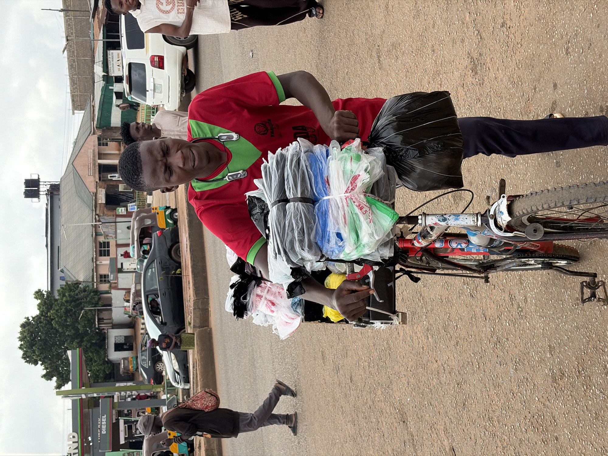 A young Black man wearing a red and green t-shirt stands on a dirt road in Gura Top, Nigeria, pushing a bicycle laden with plastic bags. He is focused on securing a large black plastic bag to the front of his bicycle. The bicycle has a red frame and is decorated with blue and white stickers. In the background, several vehicles, including cars and a white SUV, are visible, suggesting a commercial or busy area. Buildings, some with signs, line the street, and a large tree is present. Another person, also Black, walks away from the viewer in the middle ground, carrying a patterned bag over their shoulder. The sky is overcast.