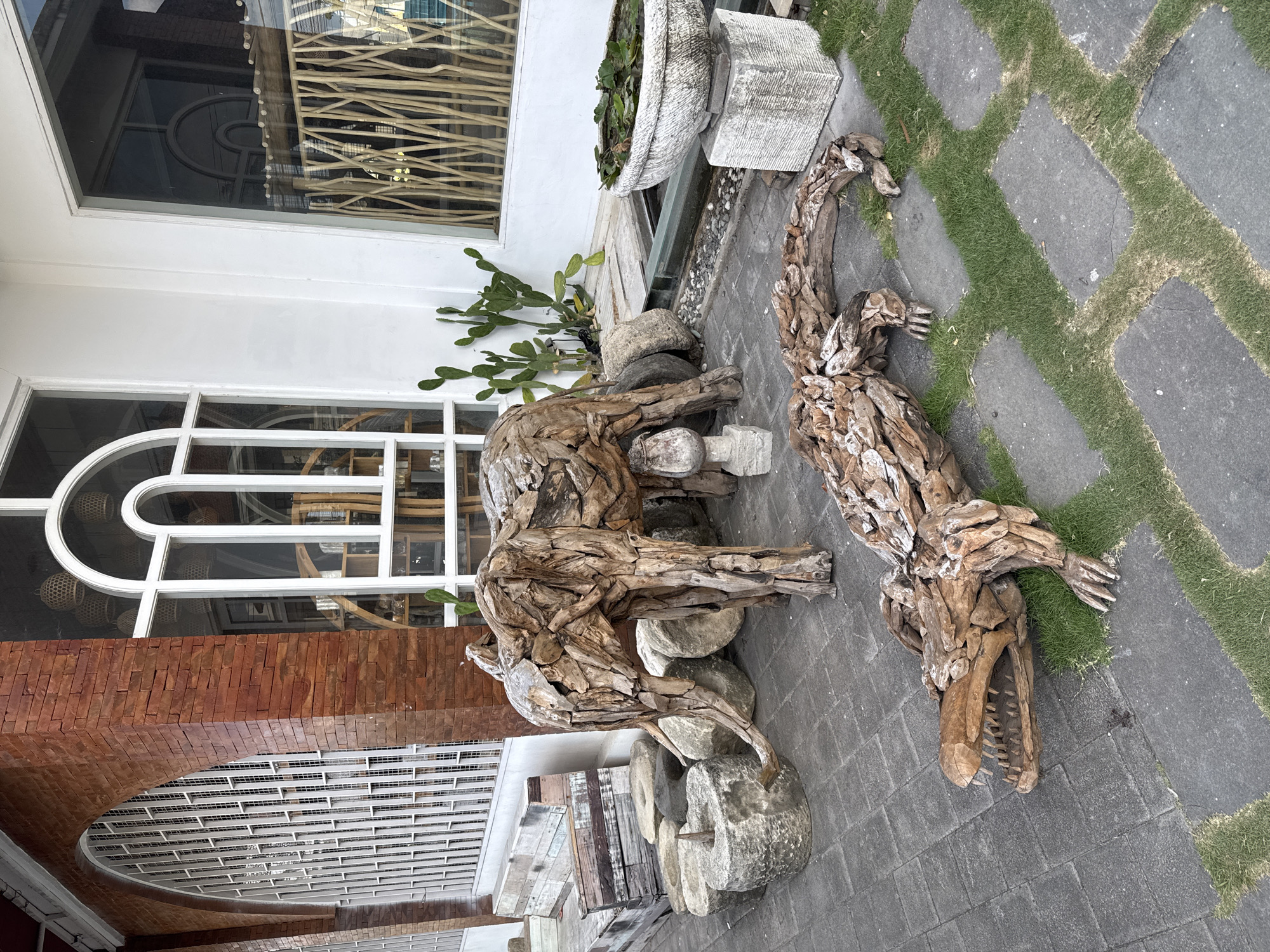 An outdoor scene in North Kuta, Indonesia, features decorative sculptures of an elephant and a crocodile, both crafted from weathered driftwood. The elephant sculpture stands to the left, with its trunk curled downwards. The crocodile sculpture is positioned in the foreground, lying on its side with its mouth open, revealing sharp teeth.
The sculptures are placed on a paved walkway with patches of green grass interspersed between the stone tiles. Behind the elephant, several large, rough-hewn stones and a stone mortar are visible. Further back, there are several green cactus plants.
The background shows a building with a brick facade and large windows. One window has a distinctive arched top with white framing and crisscrossing bars. Reflections in the window suggest an indoor space with decorative elements like hanging lamps and shelving units. A pond with water and floating leaves is situated to the right of the elephant sculpture, in front of a white wall. The overall setting appears to be an outdoor area of a commercial establishment or a decorative garden.