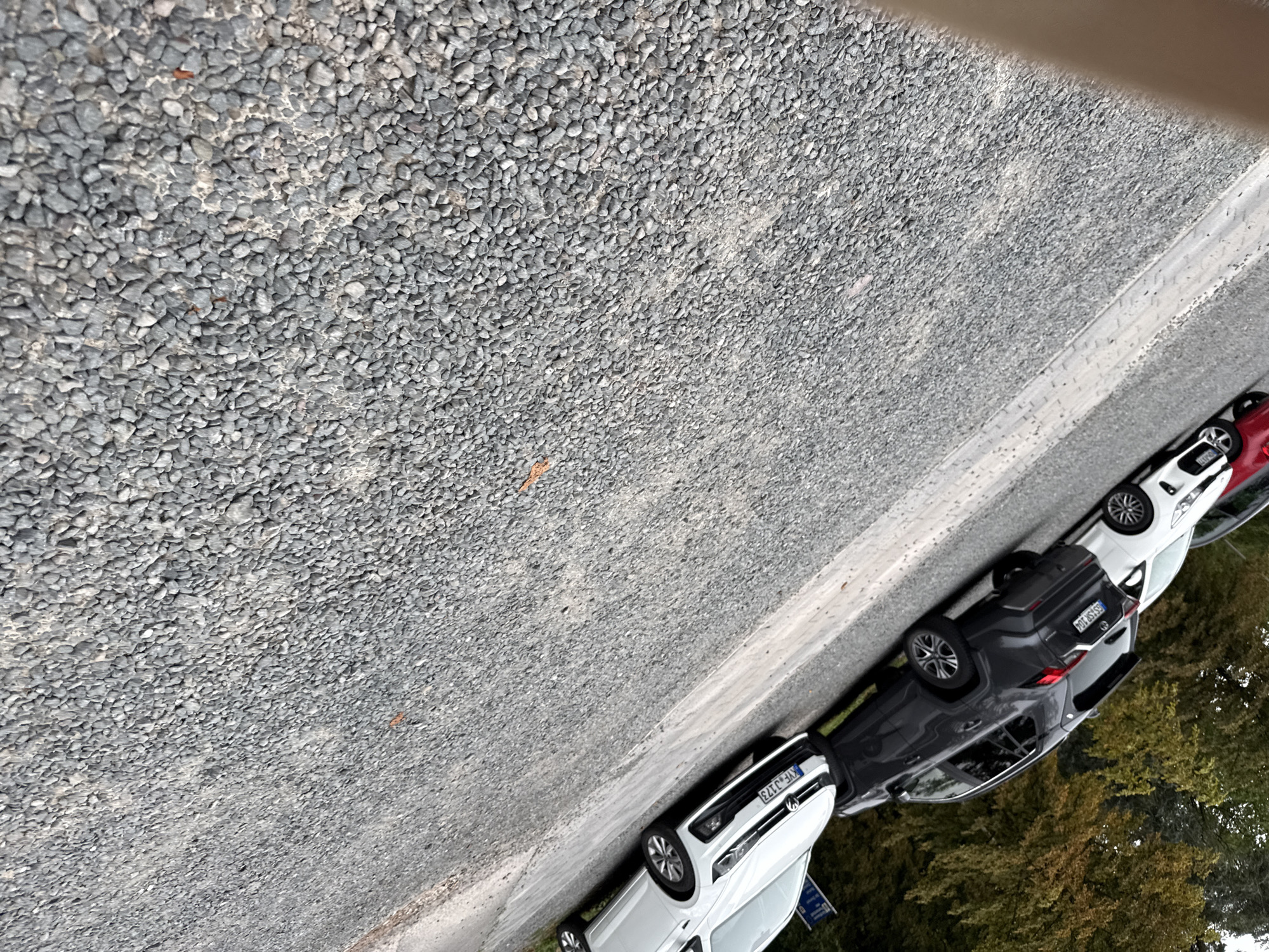 The image displays a line of parked cars alongside a gravel path. The cars appear to be in a parking area, possibly on the outskirts of Nordhausen, Germany. The terrain is predominantly covered in small, grey stones, typical of an unpaved road or parking lot. A few trees with autumnal foliage are visible to the left, suggesting the season might be fall. The cars are of various colors, including white and dark gray. One license plate is partially visible and reads "XE-JJ73". The overall scene depicts a static parking situation with no human activity or dynamic events.