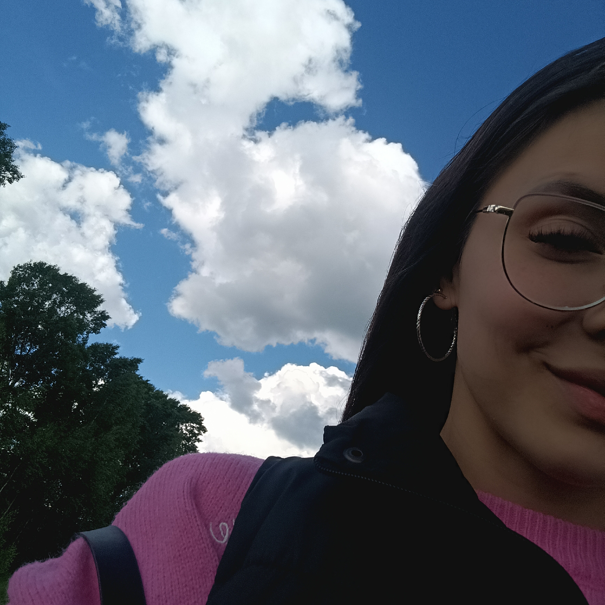 A young woman poses in an outdoor setting, presumably in Unknown, Unknown, as no specific location is provided. The right half of her face is visible, adorned with eyeglasses and a hoop earring. She wears a pink sweater under a black vest. The background features a blue sky with puffy white clouds and the dark green foliage of trees.