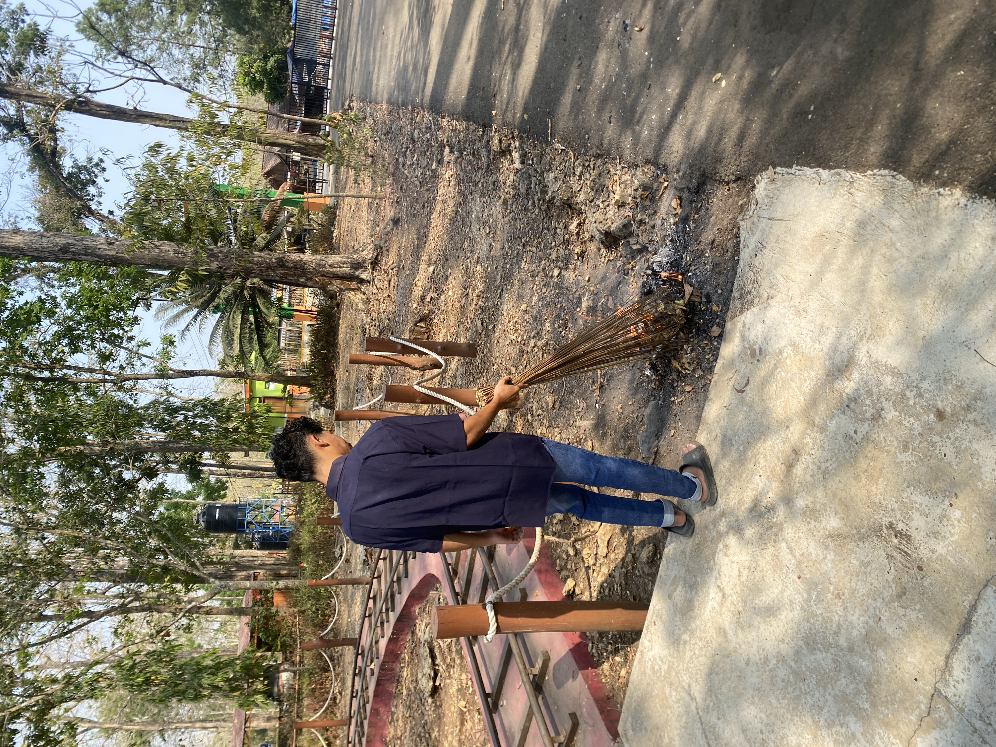 A person wearing a dark blue short-sleeved shirt and blue jeans is sweeping dry leaves into a small fire with a broom made of twigs. The person is standing on a concrete path next to a dirt area with scattered leaves and debris. Wooden posts with rope are visible in the background, bordering a path or walkway. Trees with sparse foliage and a water tank on a metal frame are also present. The ground is dry and dusty, and the scene appears to be outdoors during the daytime.