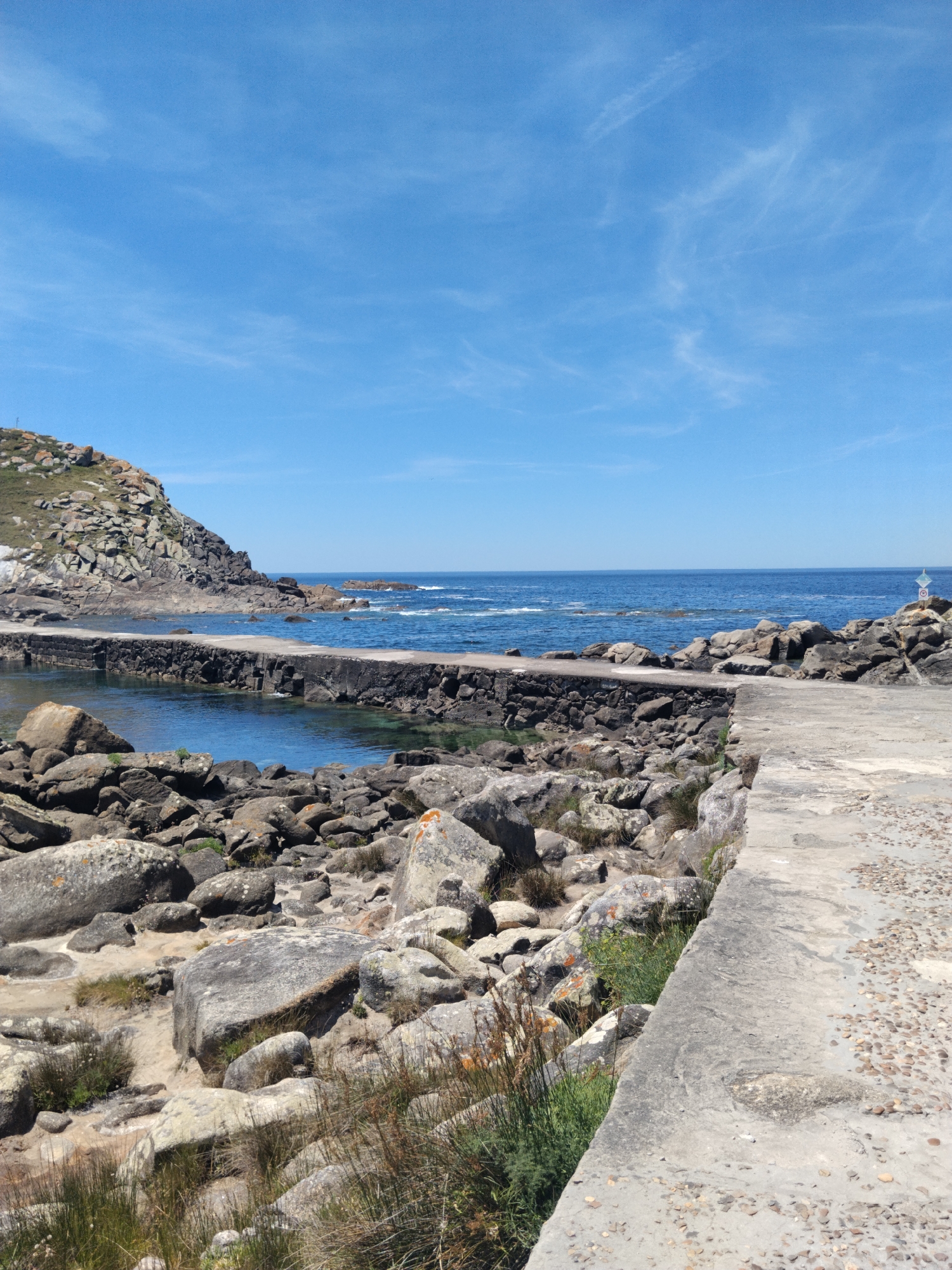 This is a scenic photograph of a coastal area in Vigo, Spain. The image features a clear blue sky with a few wispy clouds, extending over a calm blue ocean. In the foreground, a rocky shoreline is visible, with large, weathered boulders scattered along the edge. A concrete pathway or pier runs along the water's edge, with a stone wall bordering a sheltered inlet. The water in the inlet appears clear and shallow. Beyond the inlet, more rocky outcrops rise, and the ocean stretches out to the horizon. The overall impression is one of a natural, tranquil coastal landscape under a bright, sunny sky.