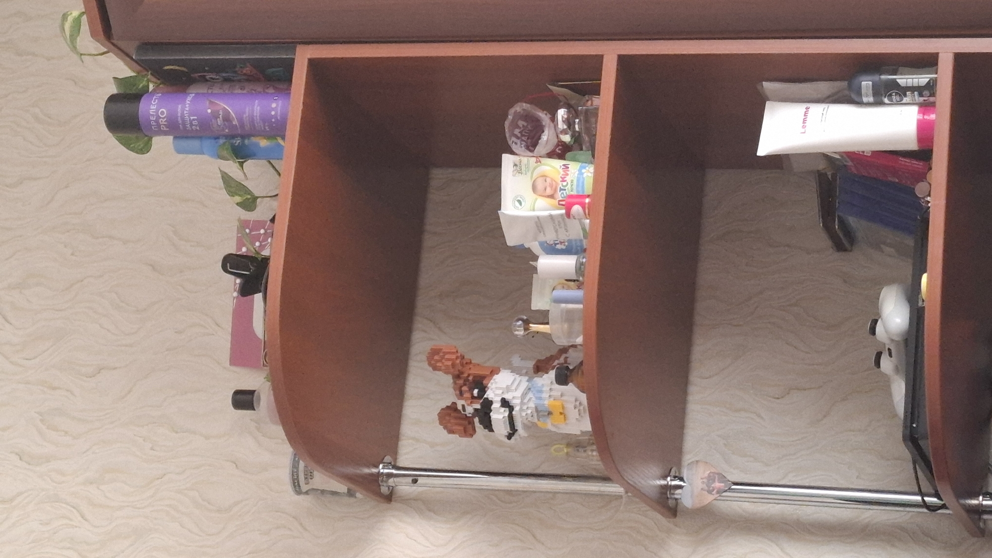 The image depicts a cluttered shelf in a domestic setting, possibly an apartment. The shelves are made of dark wood and are supported by chrome poles. On the top shelf, several items are visible including a purple hairspray can, a blue bottle, a pink box, a black binder clip, and a small plant with green leaves. The middle shelf contains a Lego-style dog figurine, a glass perfume bottle, a white cylindrical object, and a white box with a picture of a baby on it. The bottom shelf holds a gaming console, two white game controllers, a laptop, a white tube with "Lemme" written on it, and a black deodorant spray can. The background is a textured wallpaper. No specific location in Shymkent, Kazakhstan can be determined from the image.