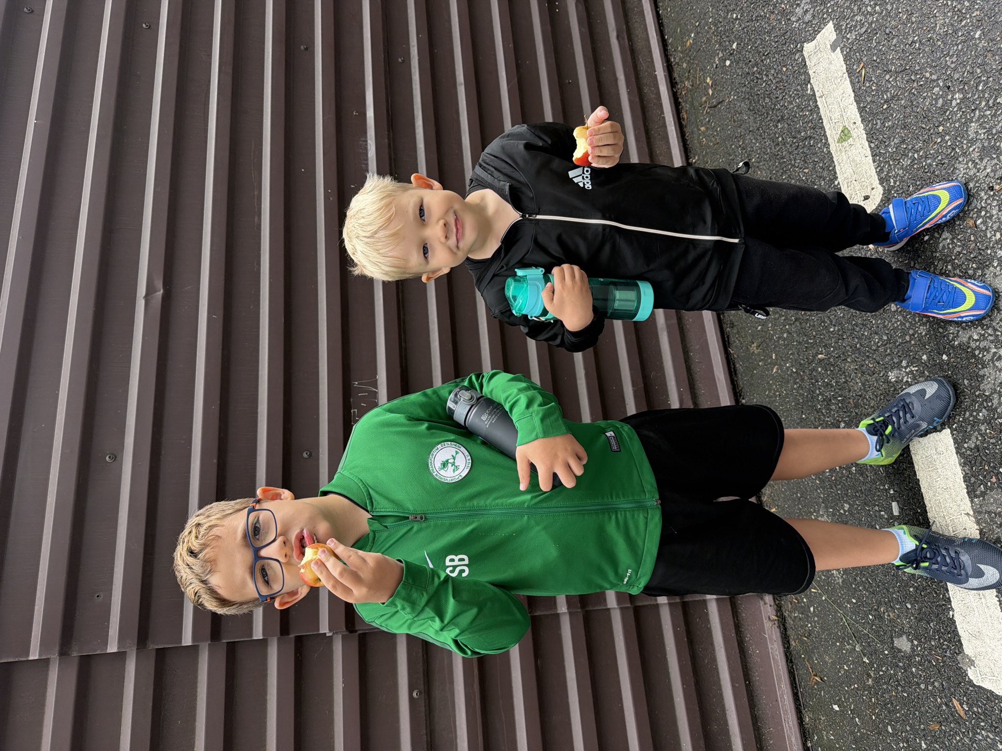Two young boys are standing side-by-side outdoors. The boy in the foreground wears glasses, a green tracksuit jacket with the letters "SB" on it, and black shorts. He is eating an apple. The other boy wears a black Adidas tracksuit and is holding a water bottle. They are standing on asphalt next to a metal wall. The location is Sochi, Russia.