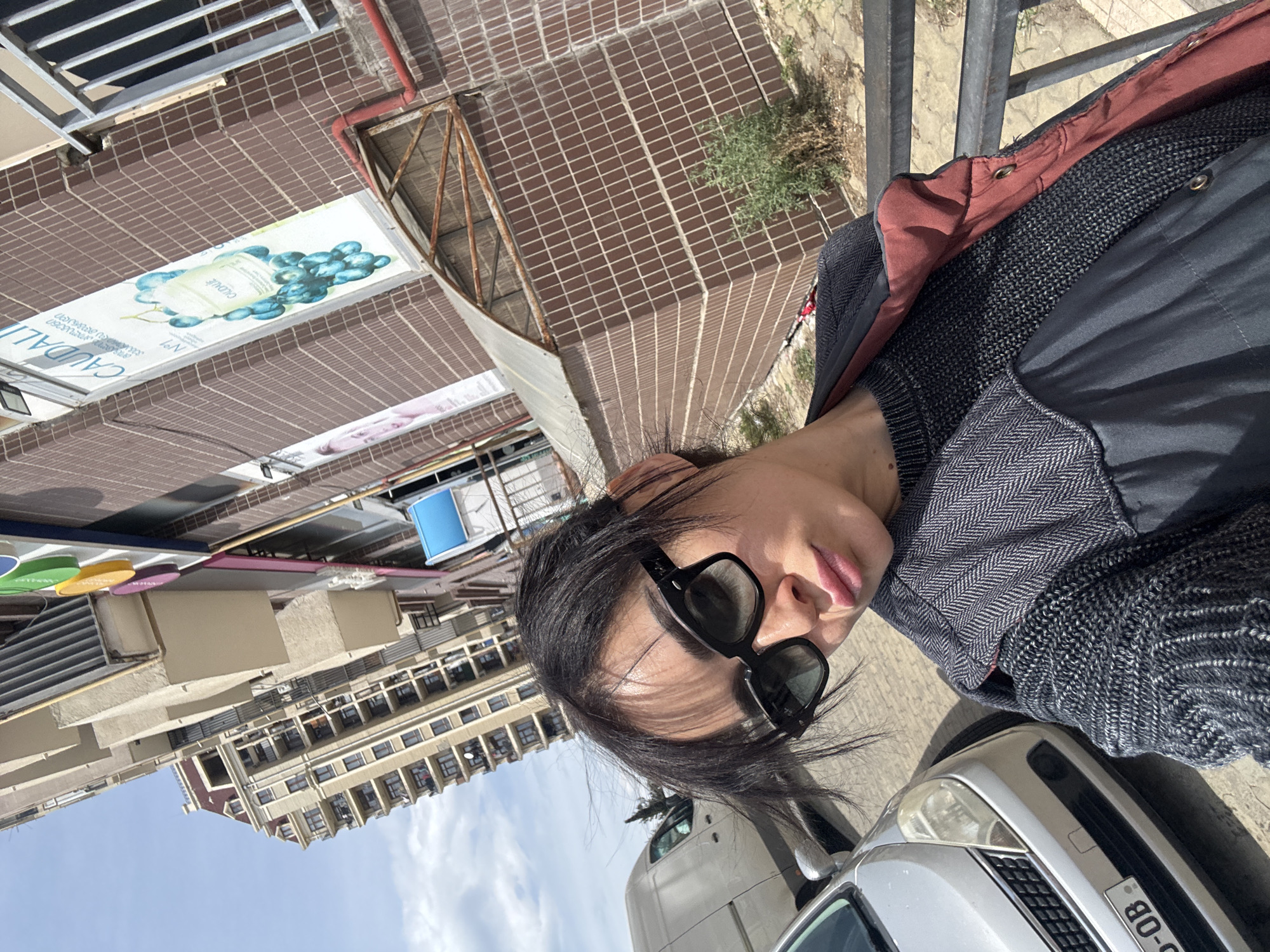 The image is a rotated outdoor selfie taken during the daytime in an urban environment, likely in Tbilisi, Georgia. The primary subject is a person, appearing to be female, captured in a close-up shot looking directly at the camera. She wears dark, stylish sunglasses, a dark jacket with a reddish-brown lining, and a gray sweater with a herringbone pattern on the collar area and a knitted texture on the main body. Her dark hair is slightly windswept.
In the foreground, a silver car is partially visible on the right, with its front bumper, grille, and headlights in view. The background reveals a street scene with several buildings under a clear, bright blue sky, indicating sunny weather. One building features a distinctive facade covered in small, dark reddish-brown tiles. There is also a metal railing and some sparse greenery visible. Further back, another multi-story building with numerous windows and balconies can be seen, alongside a partial view of a white truck or van.
The activity depicted is the person posing for a photograph.
Notable details include the strong sunlight creating distinct shadows, and the unique architectural style of the surrounding buildings. Visible text includes "CAUDALIE" and "Nº1" on a sign attached to the tiled building, which also features an image of grapes and a cosmetic bottle. Part of a vehicle license plate, reading "0B-0", is visible on the silver car. The image itself is oriented 90 degrees counter-clockwise.