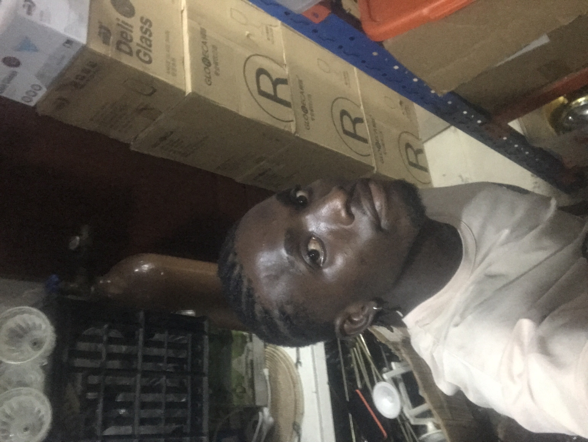 The image, oriented on its side, captures a man looking directly at the viewer within what appears to be a cluttered storage room or back area. The man, with dark skin, short dark hair, and some facial hair, is positioned on the right side of the frame, wearing a light-colored, possibly white or light grey, long-sleeved shirt. His expression is neutral as he engages with the camera.
The background is dominated by stacked cardboard boxes, forming shelves or storage units. Visible text on these boxes includes "Deli Glass" on one, and a large 'R' within a circle logo, along with "GLOOCA" or "GLOOCARE", on several others. One box also displays "000". These labels suggest the storage of commercial goods, possibly glassware.
A blue metal shelving unit is visible, holding an orange plastic container or tray. Below the boxes, a dark, grid-patterned plastic crate is present. Further in the background, an amber-colored bottle or cylinder lies horizontally. To the lower left, a stack of white, translucent, bowl-shaped or fan-like items can be seen. On the far right, near the man's shoulder, thin rods or pipes and a small white conical object are discernible. The overall lighting is dim, indicating an indoor setting likely illuminated by artificial light.