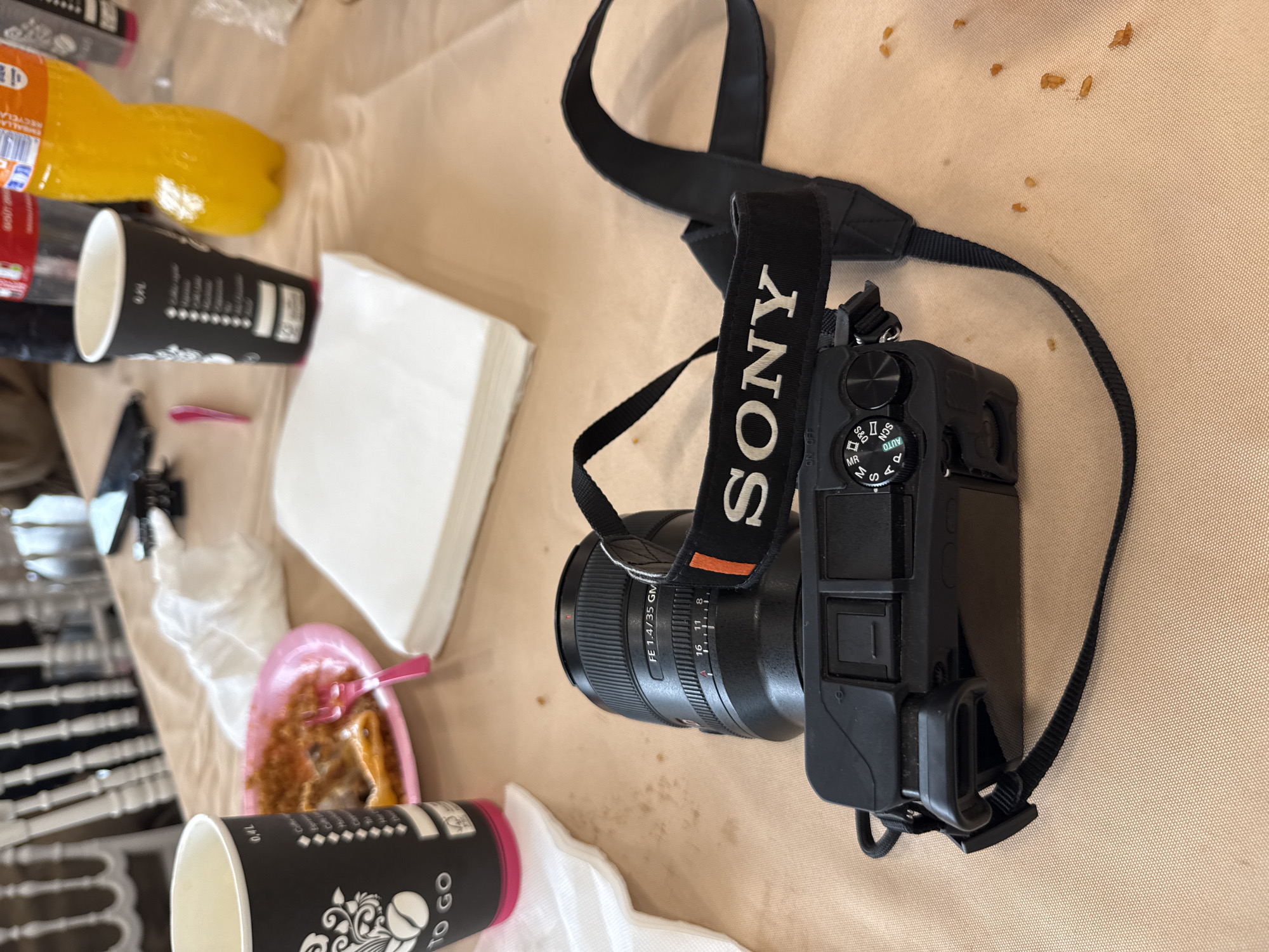 On a beige tablecloth, a Sony camera with a black strap sits amongst various items. There is a pink plate with what appears to be food on it, along with a pink plastic fork. A stack of white napkins is also visible. Black coffee cups with "TO GO" printed on them and a bottle of orange juice can be seen. The setting suggests an indoor dining or meeting area.
