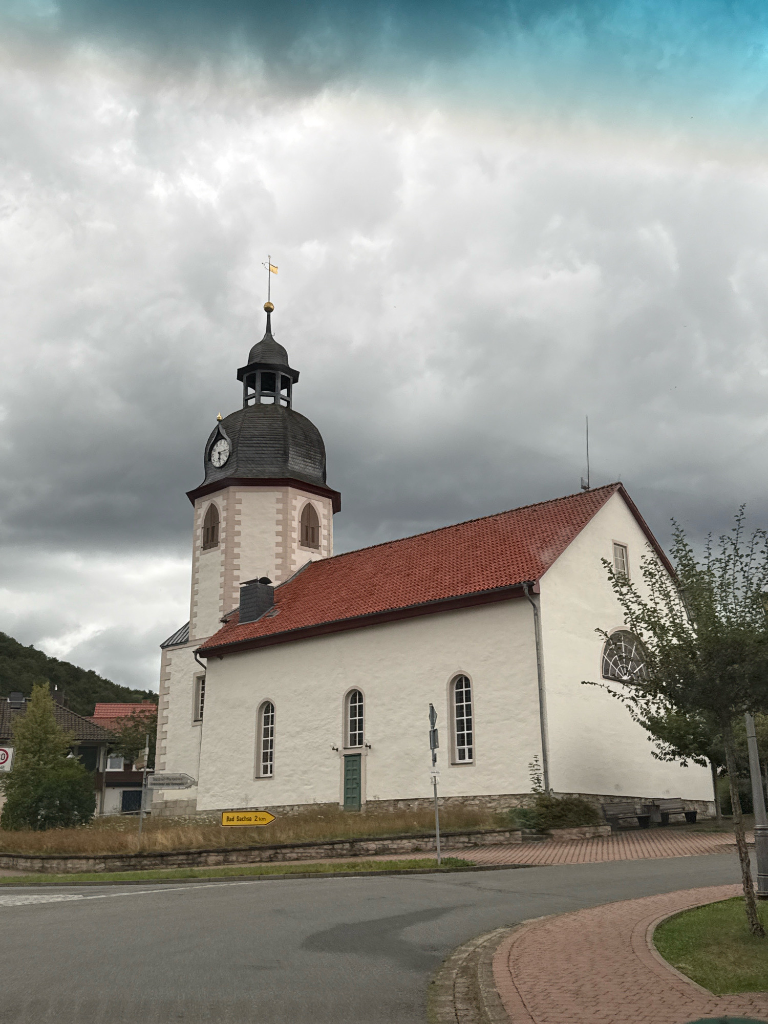 A white church with a red tile roof and a black steeple is pictured in Bad Sachsa, Germany. The church has a clock on its steeple and arched windows. The sky is cloudy and appears to be either dawn or dusk. There is a road in the foreground with a yellow directional sign indicating "Bad Sachsa 2 km". Trees and hills are visible in the background.