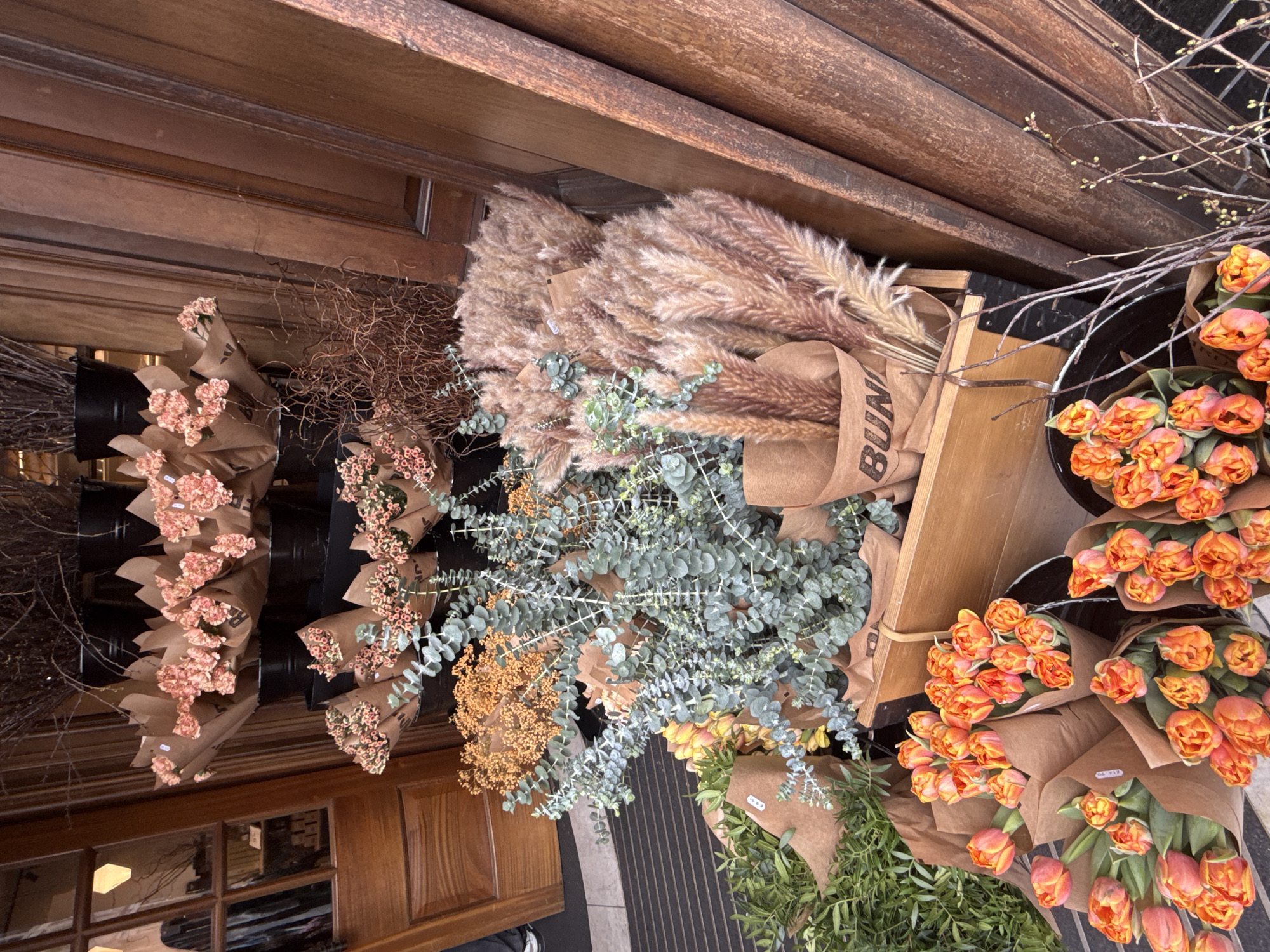 The image shows a display of various cut flowers and ornamental grasses in front of a shop, likely a florist, in London, United Kingdom. The shopfront appears to be traditional, with dark wooden panels and a framed window. There are several types of flowers, including peach carnations, orange tulips, and dried flowers, each bundled and wrapped in brown paper. Ornamental grasses are also present, including pampas grass and eucalyptus, bundled and displayed in a wooden crate. The flowers are arranged in black buckets, some positioned directly on the street pavement. The pavement itself is a dark gray with a grooved pattern. Part of a pedestrian's legs can be seen in the lower center of the image. The overall scene suggests an outdoor flower stall or an extension of a florist shop onto the sidewalk.