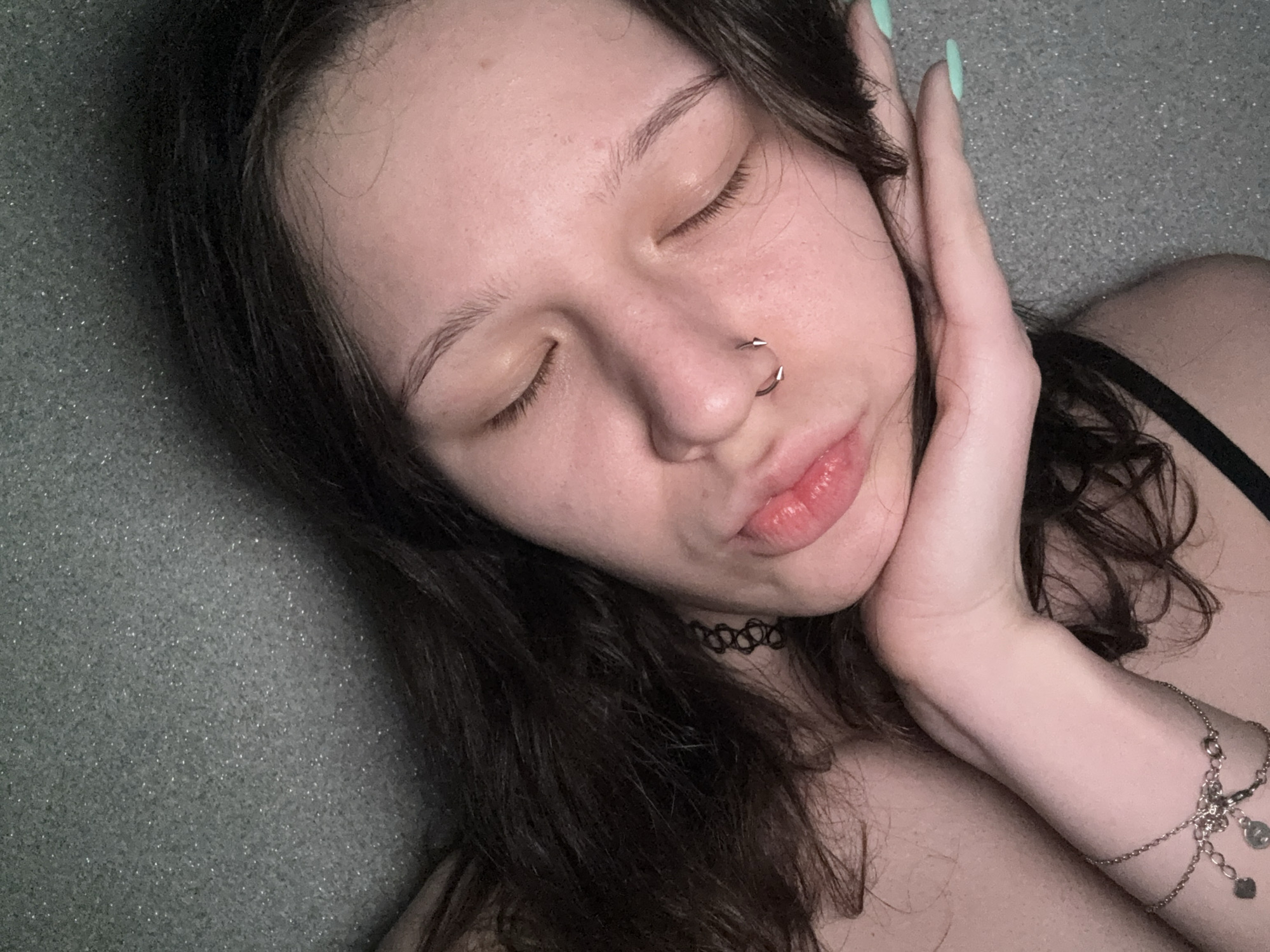 A close-up, angled portrait features a young individual, likely female, with long dark hair, against a textured, dark grey background that appears to be fabric or a wall. The person's eyes are closed, and their head is tilted to the right, suggesting a relaxed or sleeping posture, or a posed moment.
One hand is gently placed against the left side of their face, with long, light green (mint-colored) nails visible. The lips are slightly parted and puckered, hinting at a soft expression or a "kissy face." A black, patterned choker necklace is worn around the neck. A septum piercing, a silver ring with small spikes, adorns the nose. On the right wrist, a gold-toned chain bracelet with multiple small charms, including what appears to be a heart, is visible. Part of a black strap, possibly from a top or bra, is visible on the right side of the frame. The lighting is soft but appears to be artificial, making the time of day indeterminate. There is no visible text.