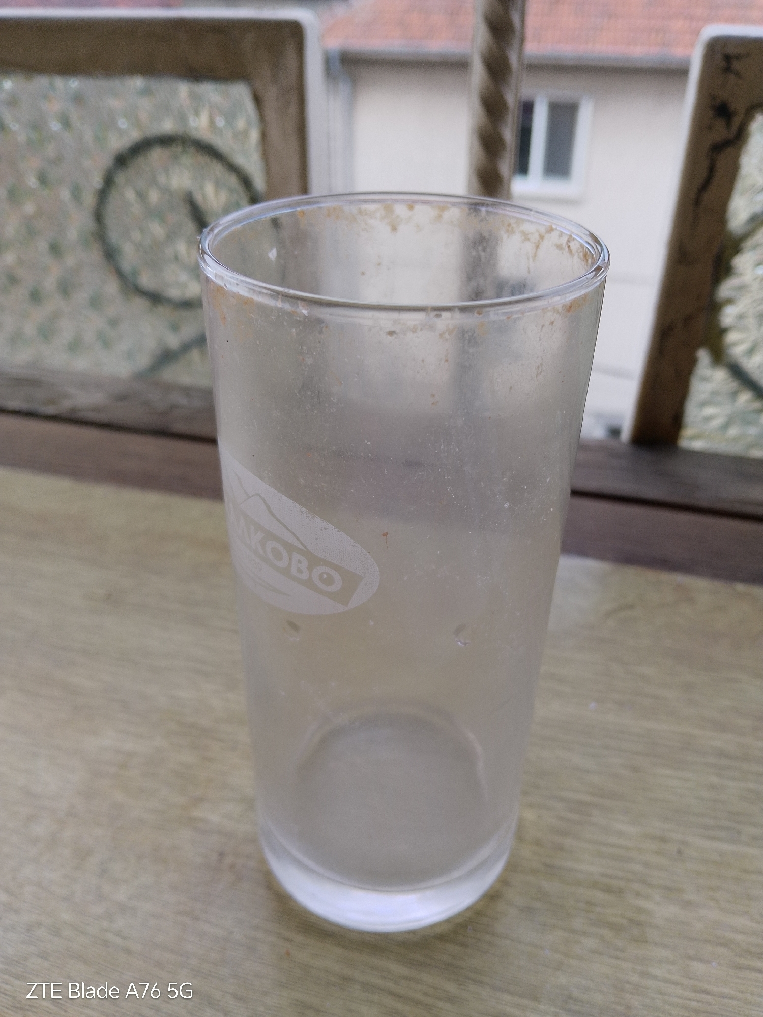 A dirty, empty glass tumbler with a faint logo is positioned on a wooden surface. The glass is frosted and appears to have dried residue on its interior and exterior. The background shows a building with a red-tiled roof and a window, suggesting an outdoor setting, possibly a balcony or patio. The image is geotagged to Stara Zagora, Bulgaria. There are no people or animals present in the scene.