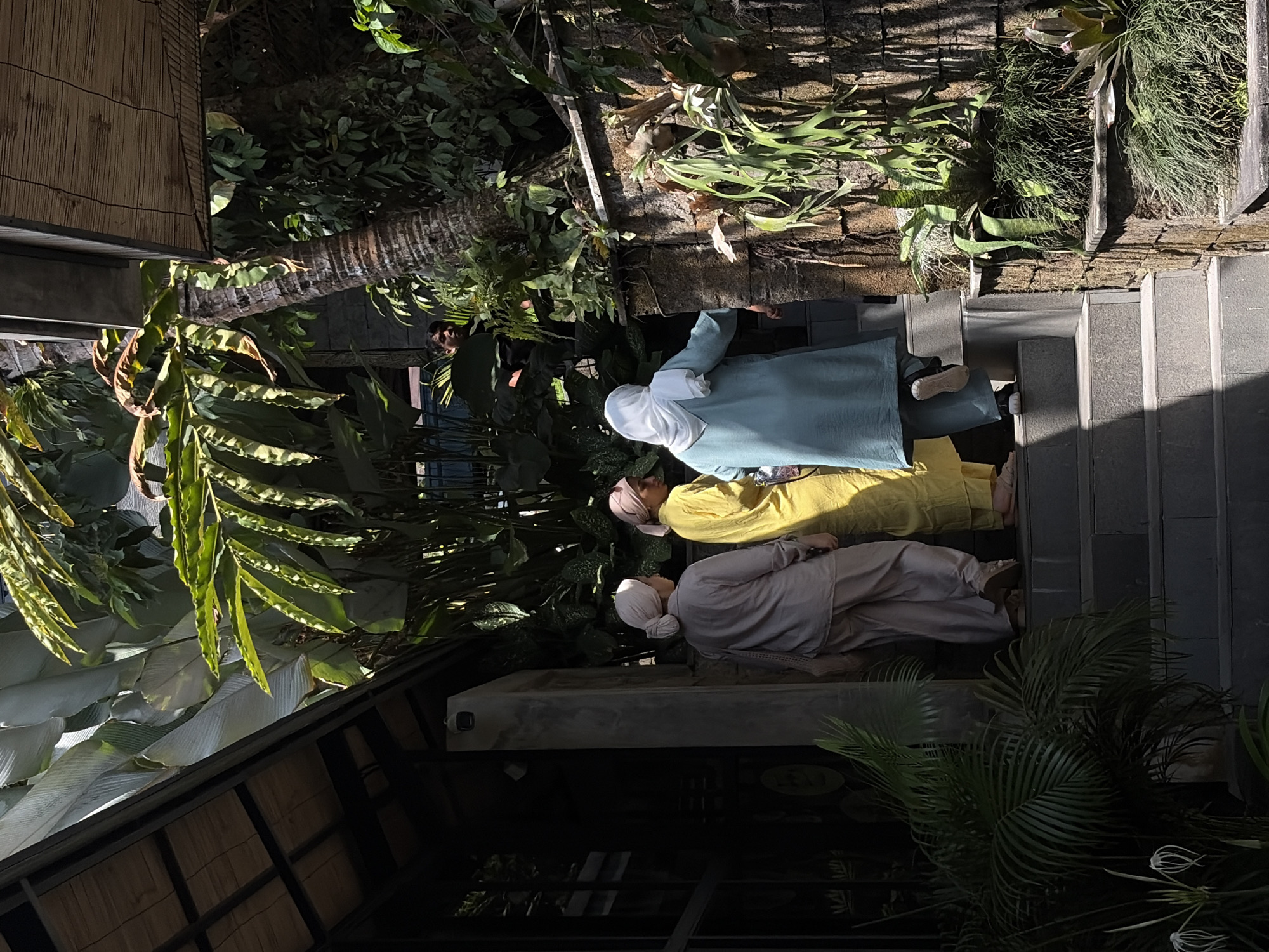 In Tegallalang, Indonesia, three individuals are ascending a set of outdoor stairs beside a lush, green vegetated wall. The person on the left wears a light brown hijab and matching loose-fitting pantsuit, carrying a beige woven bag over their shoulder. To their right, a person in a bright yellow hijab and matching pants stands, and to their right, a person in a light blue hijab and matching pants also ascends the stairs. Behind them, a man is partially visible, looking out from behind dense foliage and a palm tree. The stairs are made of dark gray stone, and the wall is constructed from rough-hewn stones adorned with various epiphytic plants like staghorn ferns. Overhead, a wooden roof structure with bamboo detailing is visible, casting shadows on the scene. The area appears to be an outdoor establishment or pathway, characterized by its natural landscaping and architectural elements.