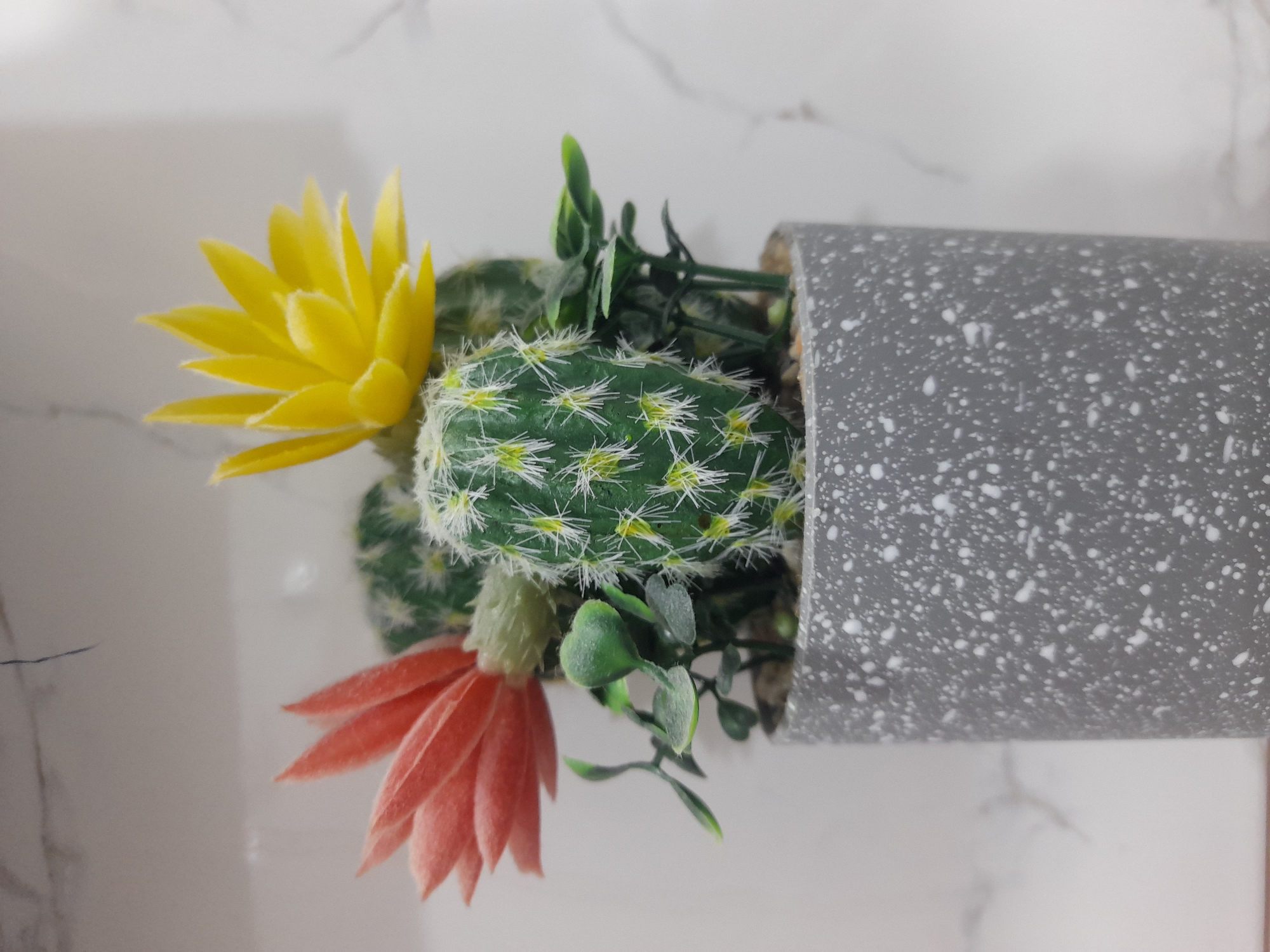 A close-up, overhead shot depicts a potted cactus with two artificial flowers. The cactus is green with white spines and is positioned in the center of a gray, speckled pot. A yellow, artificial flower is on the left side of the cactus, and an orange, artificial flower is on the lower left side of the cactus. Green leaves surround the base of the cactus. The pot is sitting on a white marble surface.